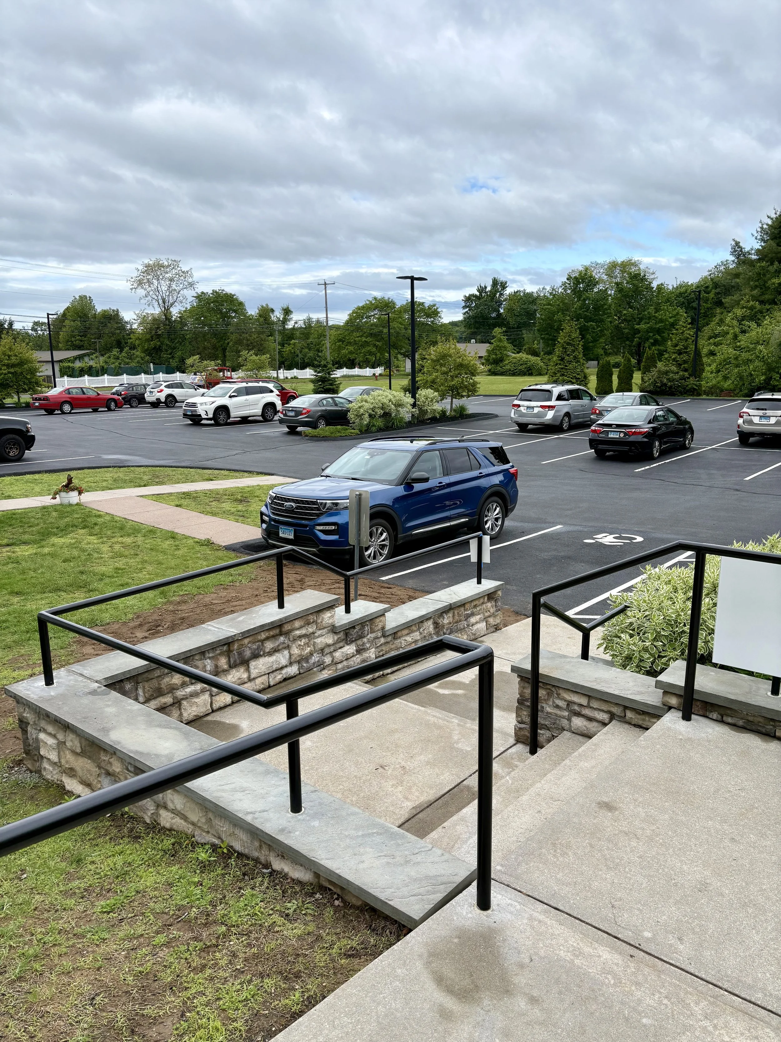 View of a parking lot with several cars and a blue SUV parked near the entrance steps of a building, with greenery and trees in the background under a cloudy sky.