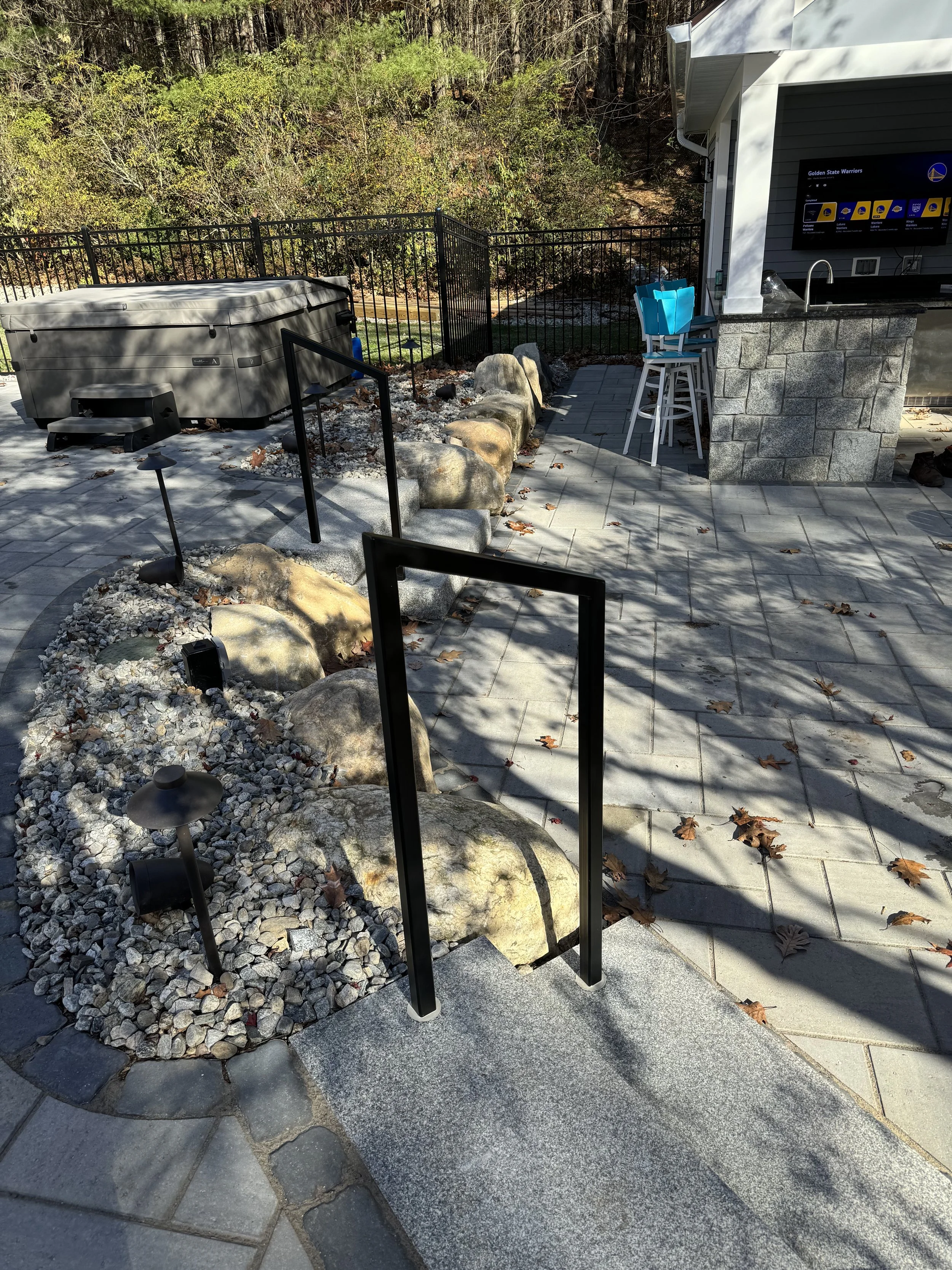 Outdoor patio with stone and gravel landscaping, large rocks, black metal handrails, a hot tub cover, a lamp, a building with a television, barstools, and miscellaneous items. There are fallen leaves on the pavement and shadows from nearby trees.