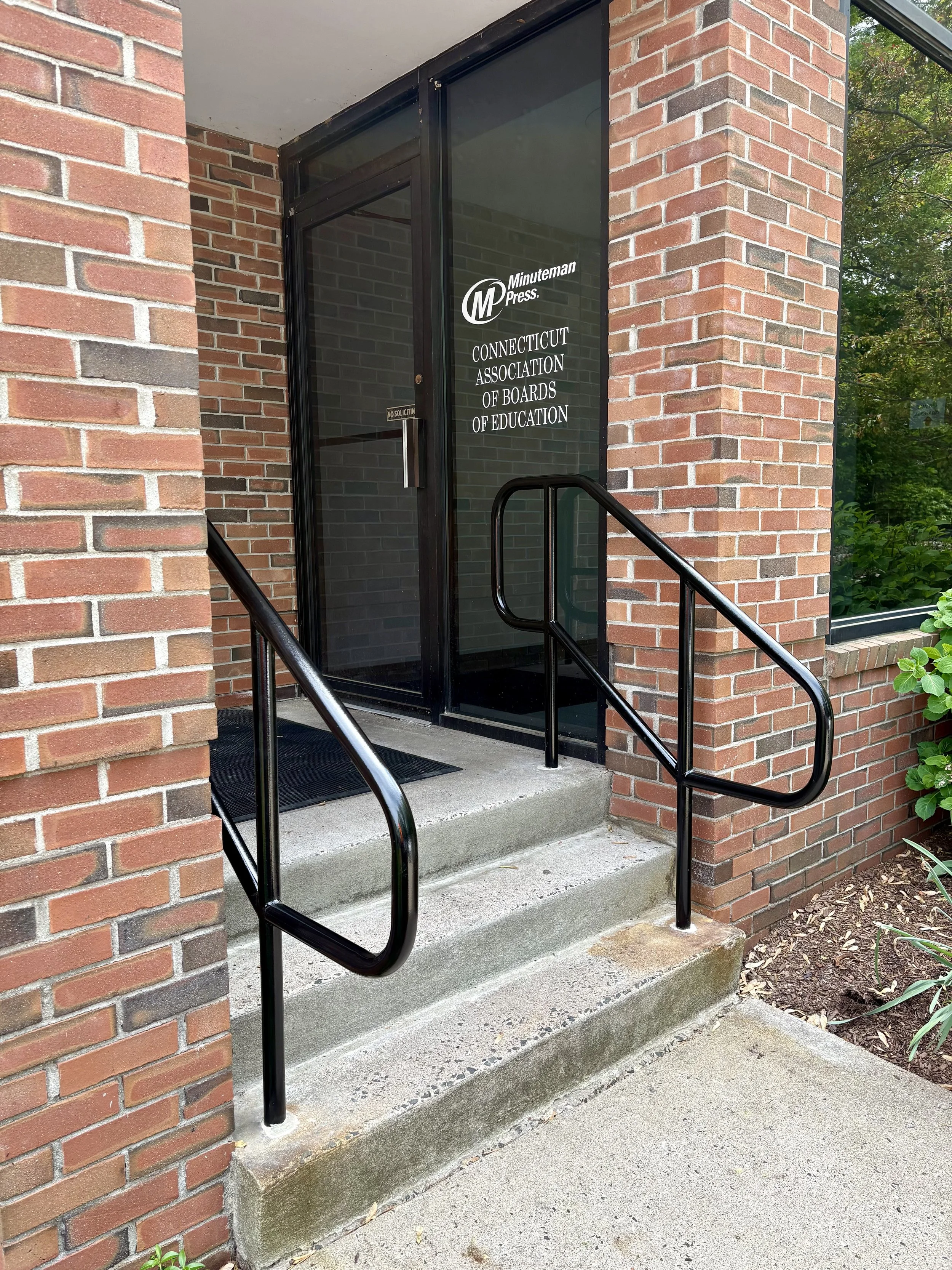 The entrance to a brick building with a glass door labeled 'Connecticut Association of Boards of Education' and 'Minuteman Press,' featuring a black metal handrail on steps leading up to the door.