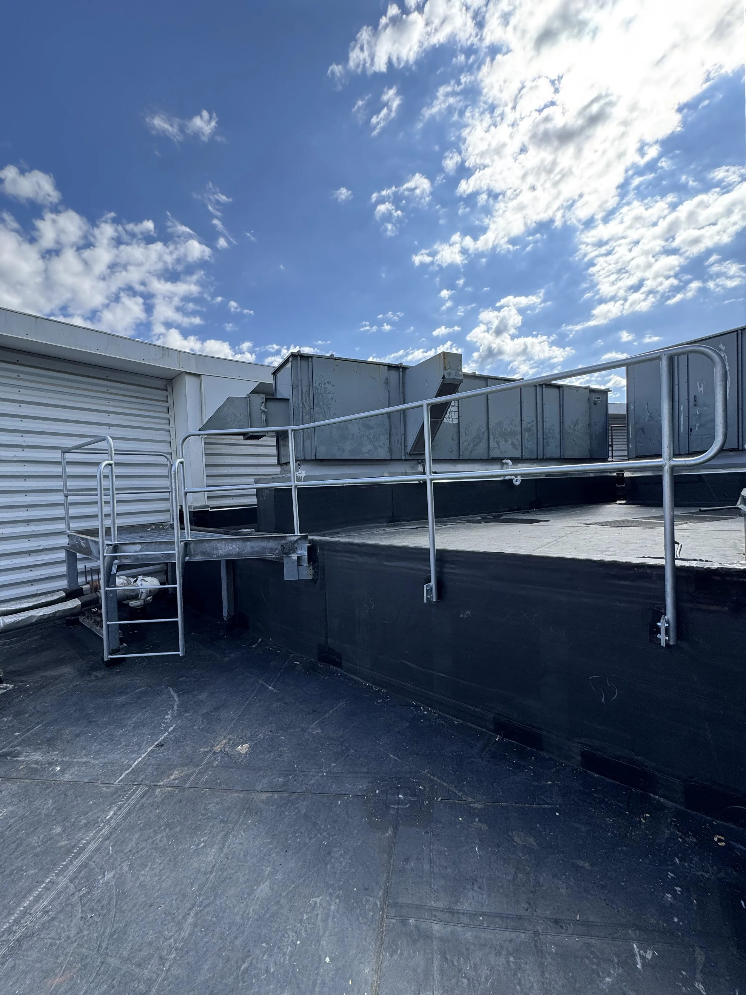 Rooftop HVAC system with metal ductwork, safety railing, and a clear blue sky with clouds.