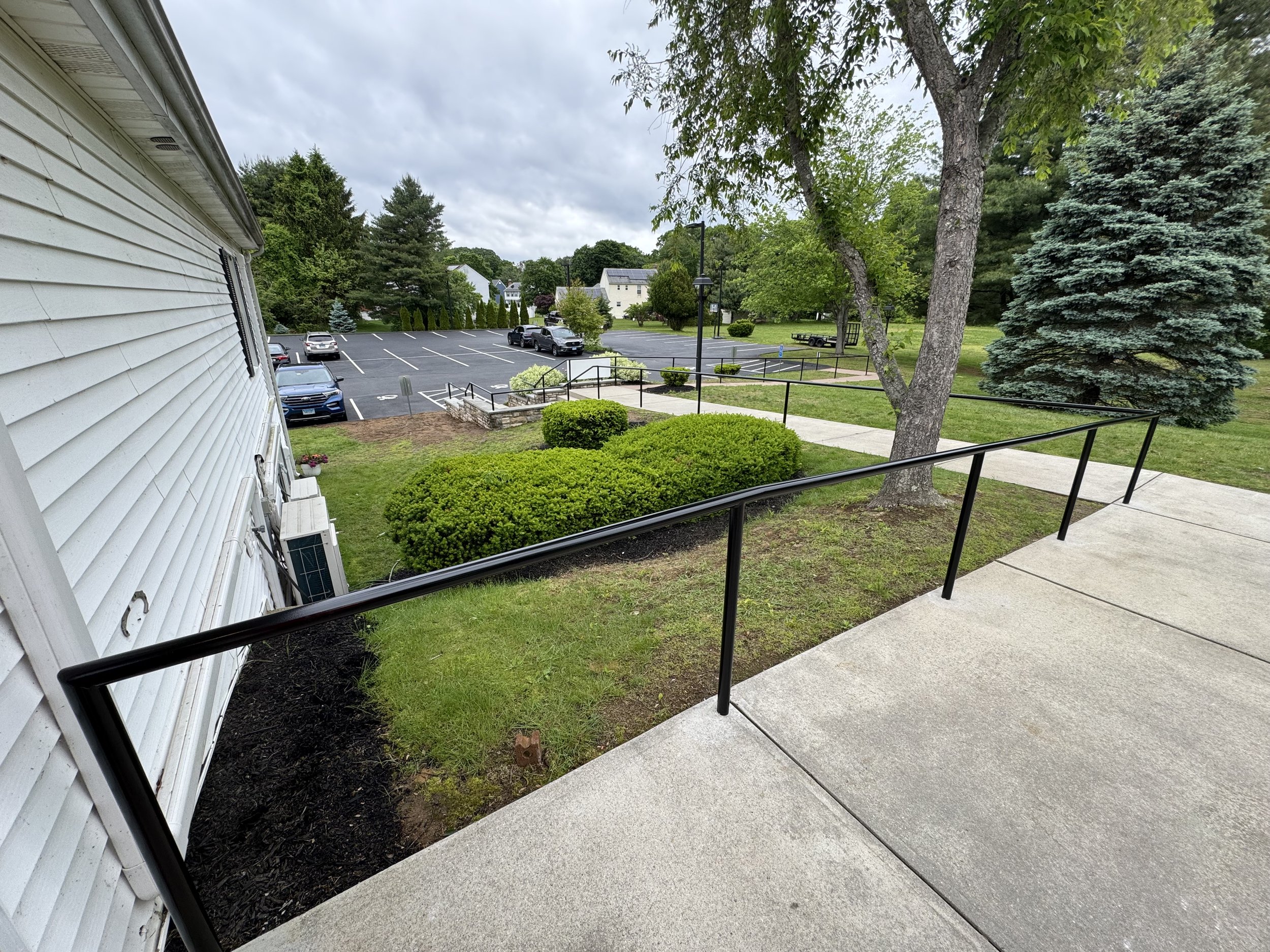 View of a parking lot and green landscape from a sidewalk with a black handrail, next to a white building with horizontal siding. Trees and shrubs are in the foreground and background.