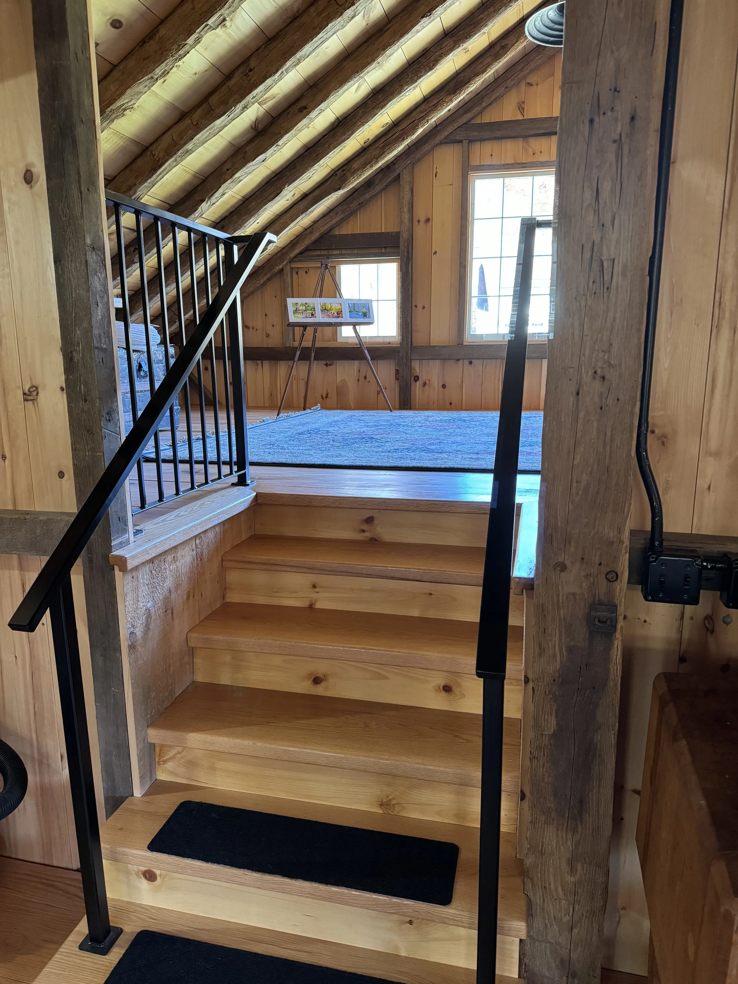 Wooden staircase with three steps and black metal handrails leading to a loft area with wood panel walls, two windows, and an easel with pictures on it.