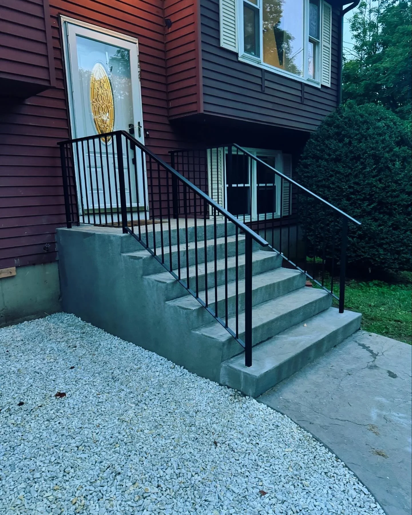 Concrete stairs with black metal railings leading up to a house entrance with a white door and golden oval glass window. The house has red siding and windows with white shutters, surrounded by green bushes and a grass yard.