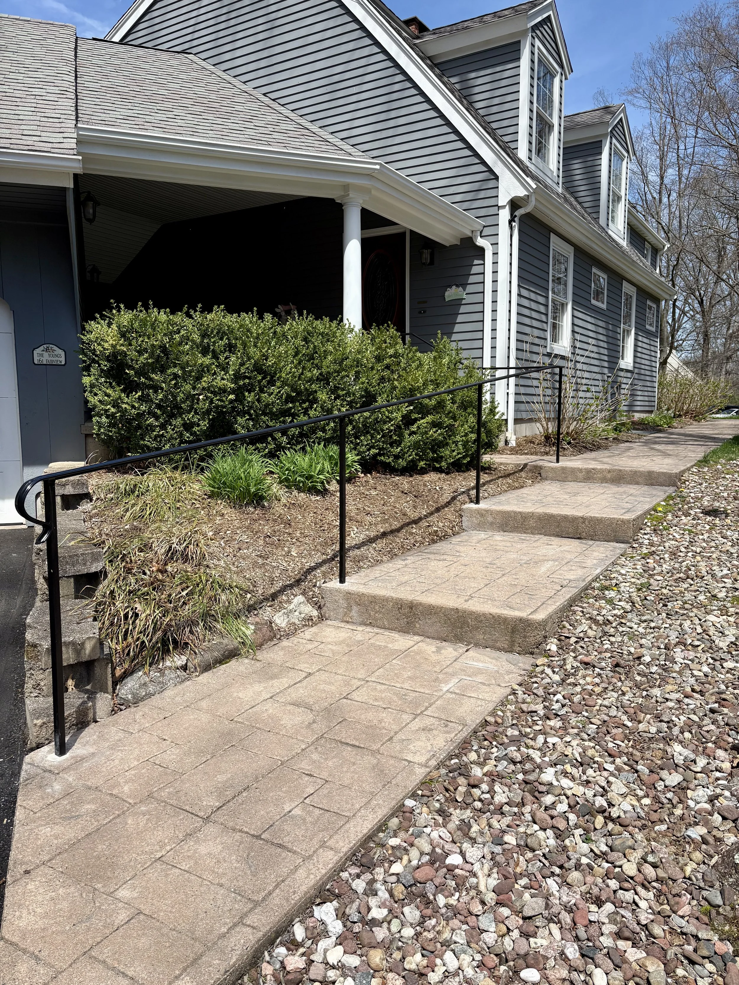 A house with grey siding and a front porch with a black metal railing. The walkway leading up to the house has several steps, with a stone-paved pathway and gravel on the side. There are bushes and small plants along the stairway and a few leafless t