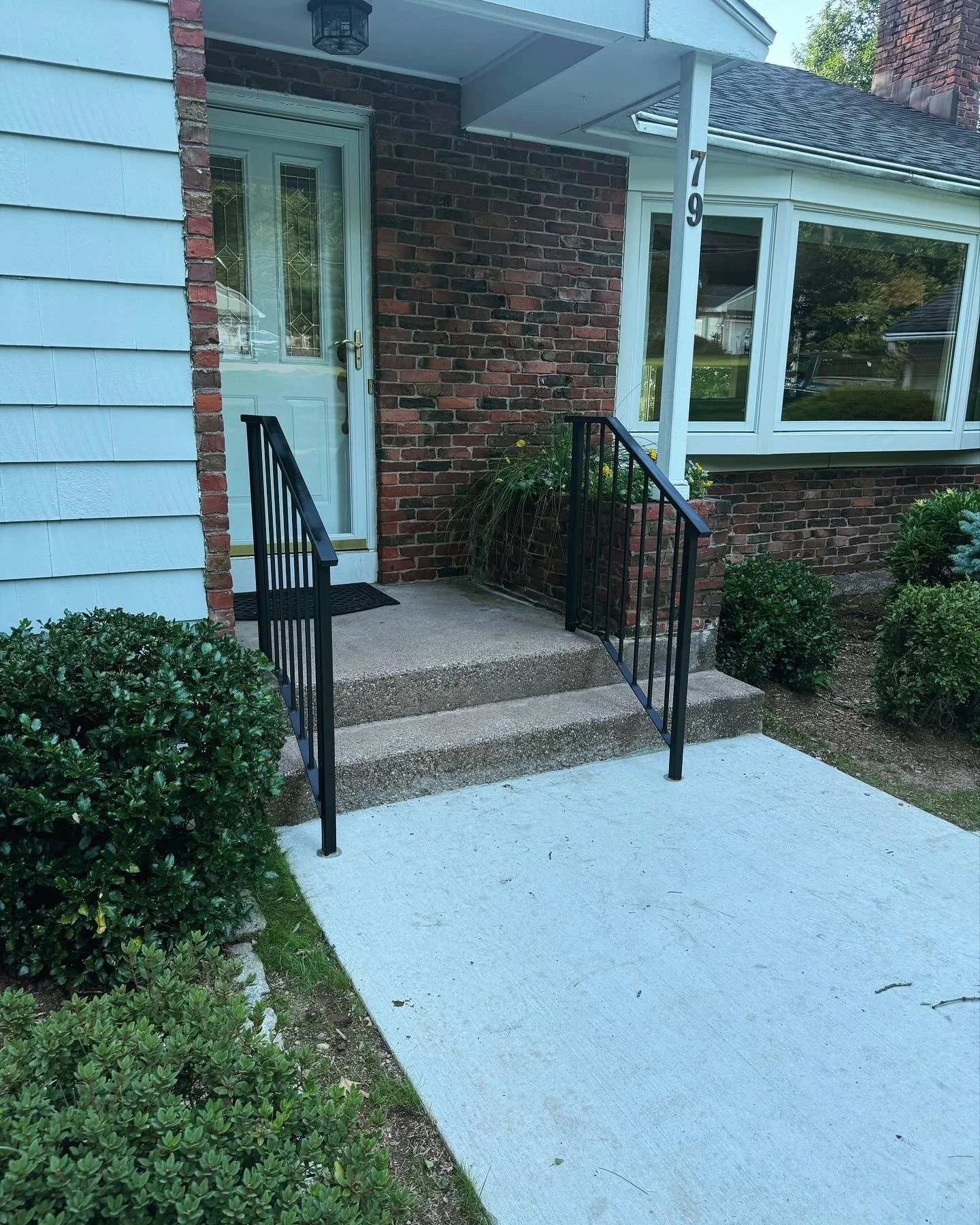 Front entrance of a house with a small front porch, concrete steps, black metal handrails, a white door with glass, and a brick and siding exterior. The house has a house number 79 and nearby shrubbery.