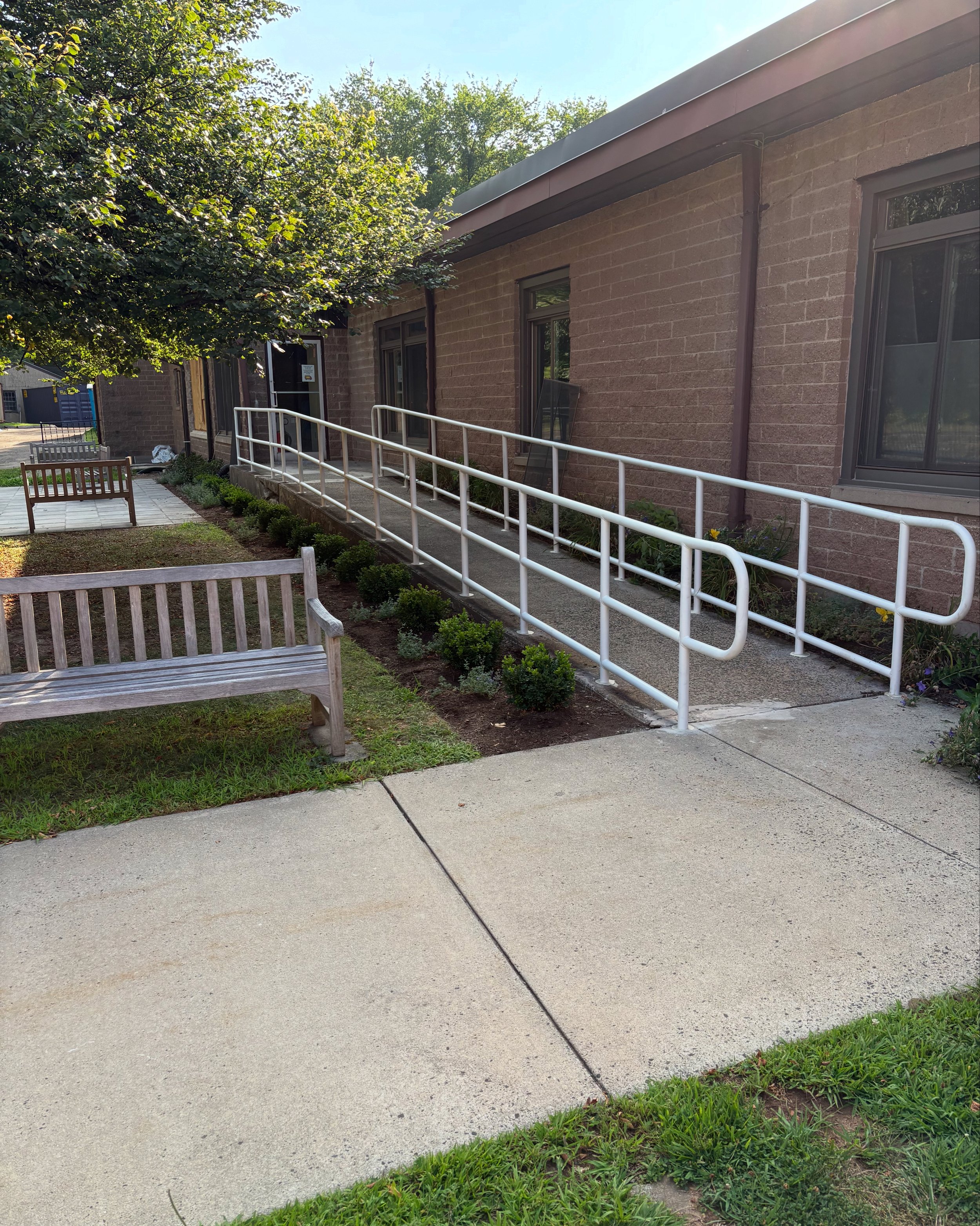 Outside view of a brick building with an accessible ramp and railing, flower beds, and benches near a sidewalk and a tree.