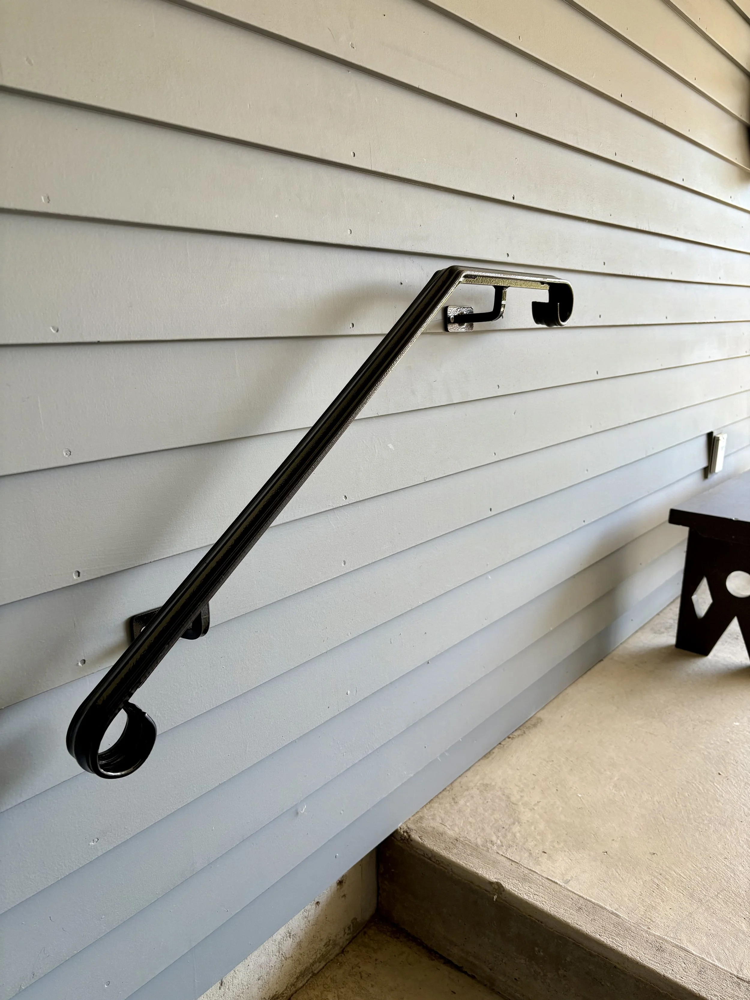 A black metal handrail attached to a light blue horizontal siding wall on a porch, with a small bench or table visible in the background.