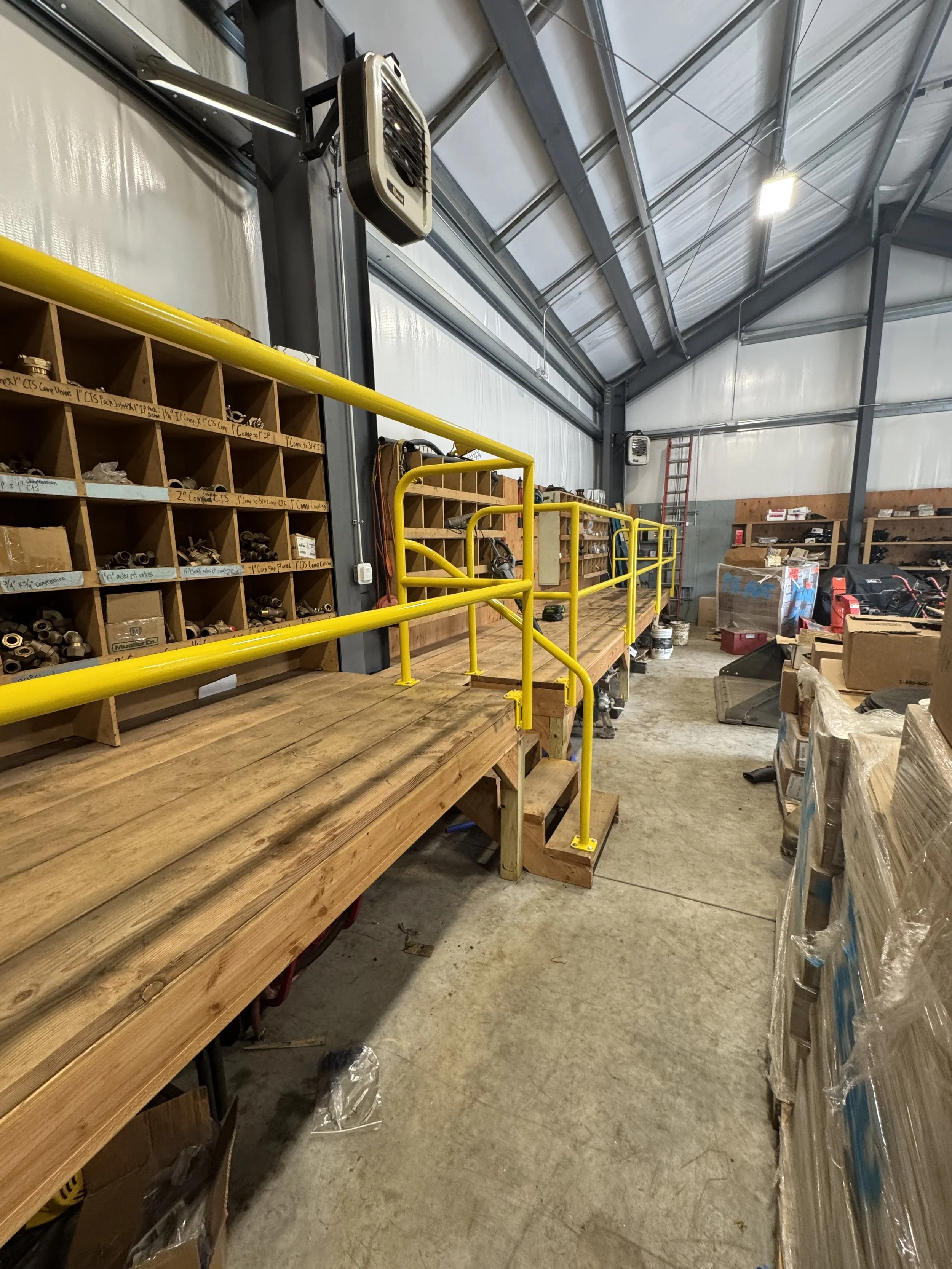 Industrial warehouse with wooden workbench, yellow safety railing, storage shelves filled with hardware parts, and organized boxes in the background.
