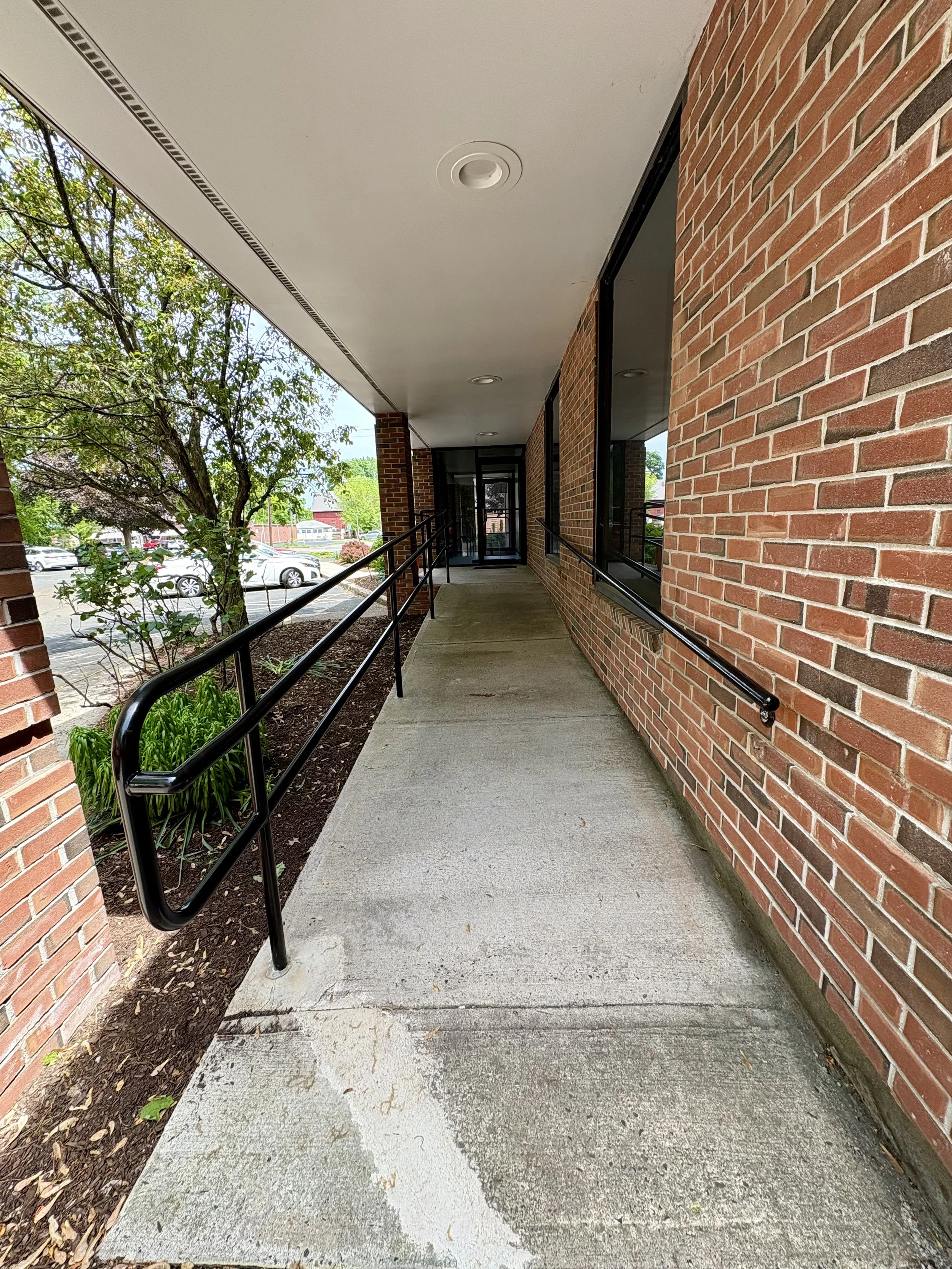 Concrete sidewalk with black metal handrails along a brick building exterior. Green trees and parked cars are visible in the background.