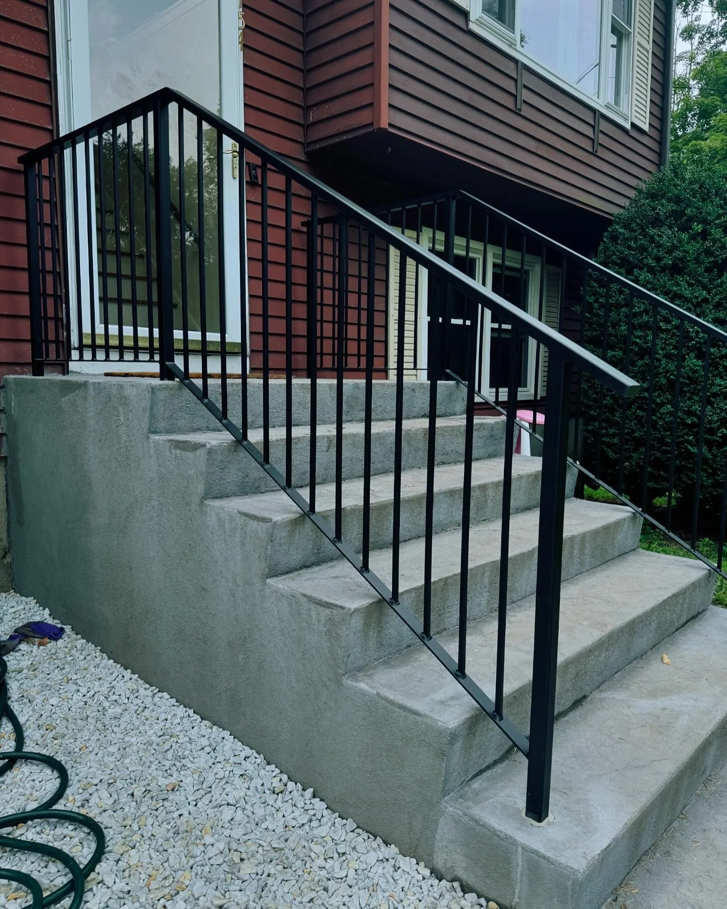 Concrete stairs leading to a house with a glass door, black metal handrail, and a bush on the right side.