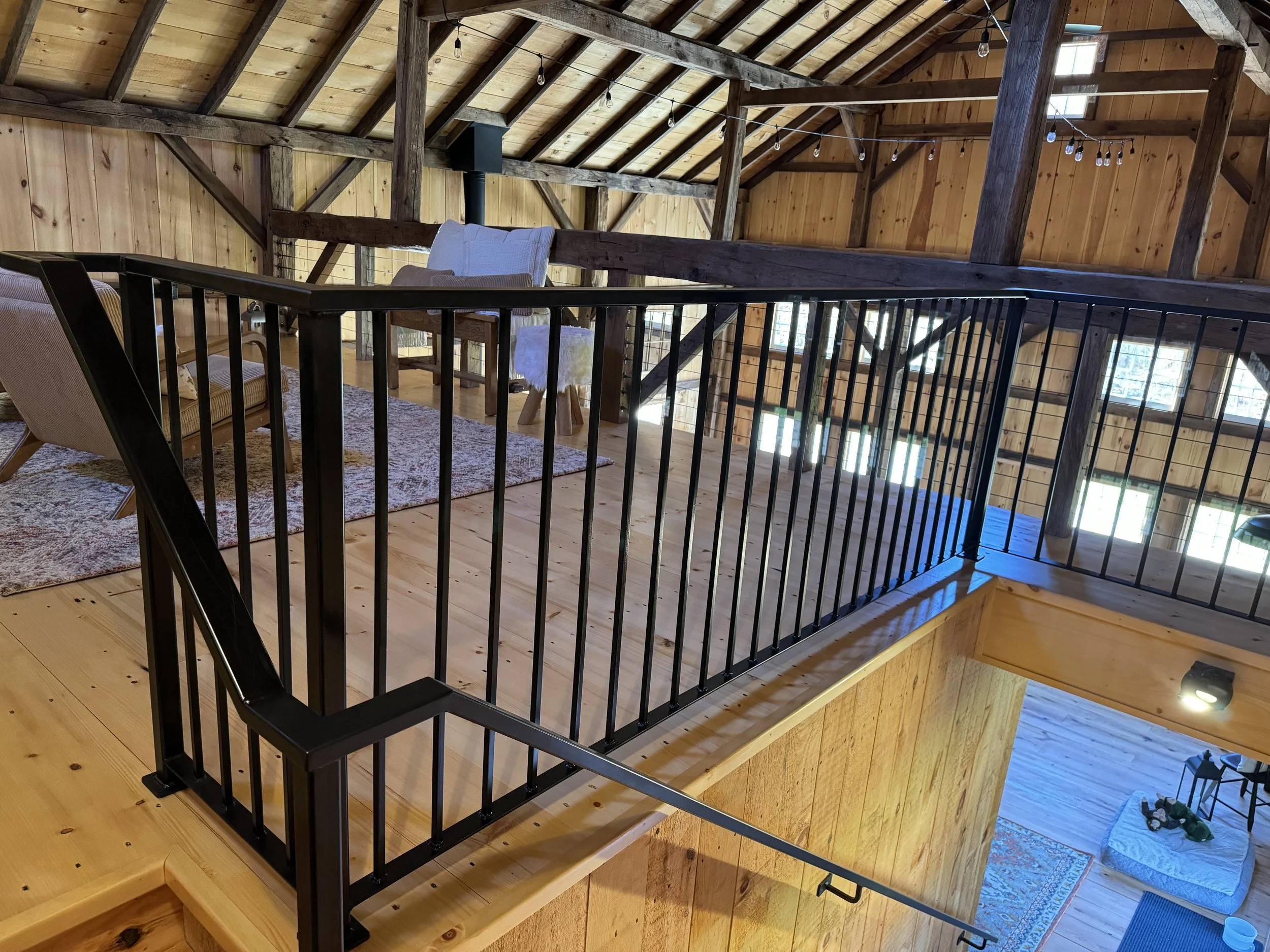 View of a cozy wooden loft area with seating, including a beige armchair and a white upholstered chair, overlooking a lower level with a bed and a patterned rug, lit by natural light from multiple windows, with string lights hanging from the ceiling.