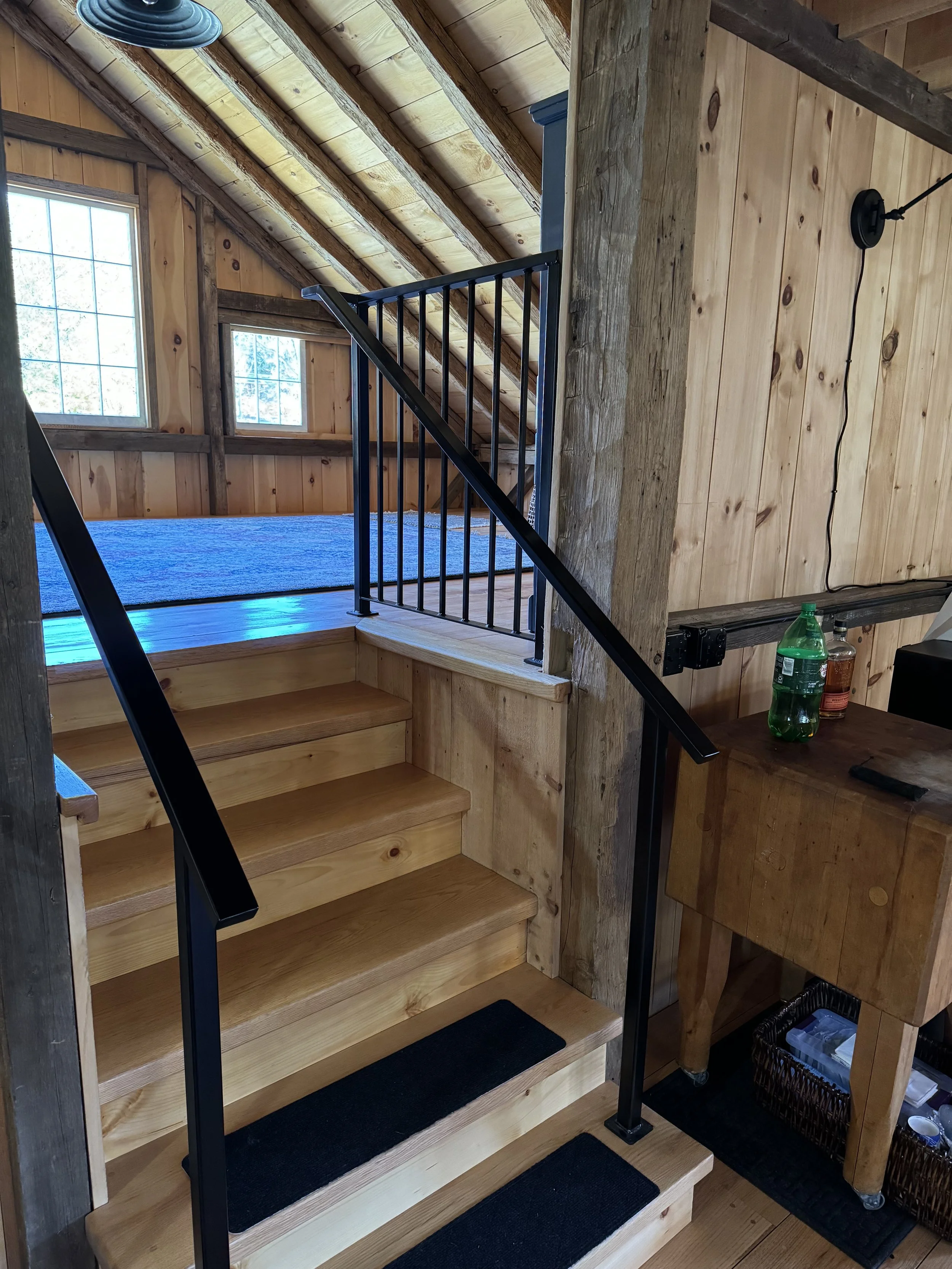 Wooden staircase with black metal railing leading to a loft area with windows, in a rustic, wood-paneled room.