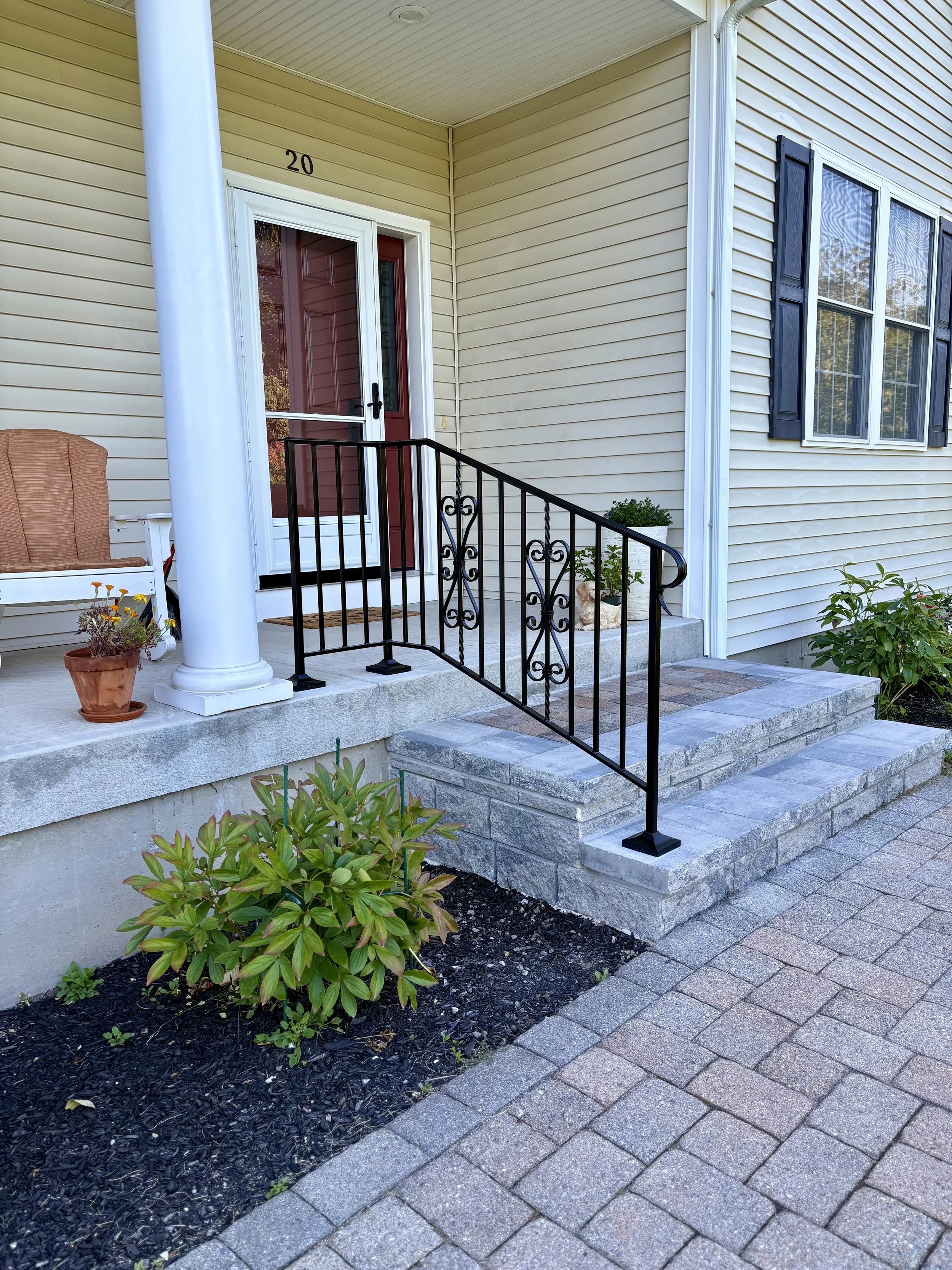 Front porch of a house with a red door, black decorative railing, two steps leading up to the door, a potted plant on the right, a small garden bed on the left with green plants, and a porch swing with cushions.