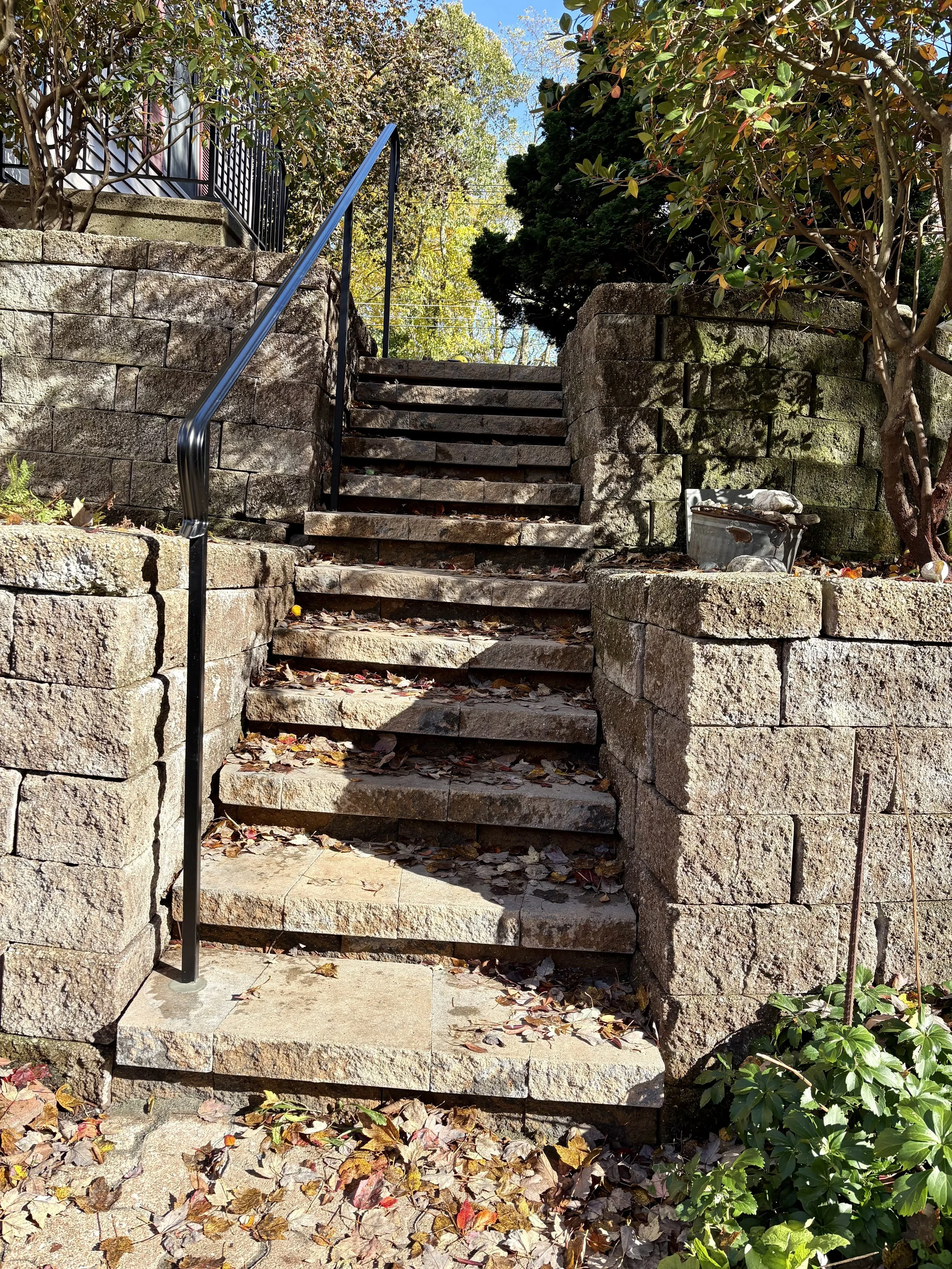 Stone staircase outdoors with a black metal handrail, surrounded by trees and bushes, with fallen leaves on the steps and ground.