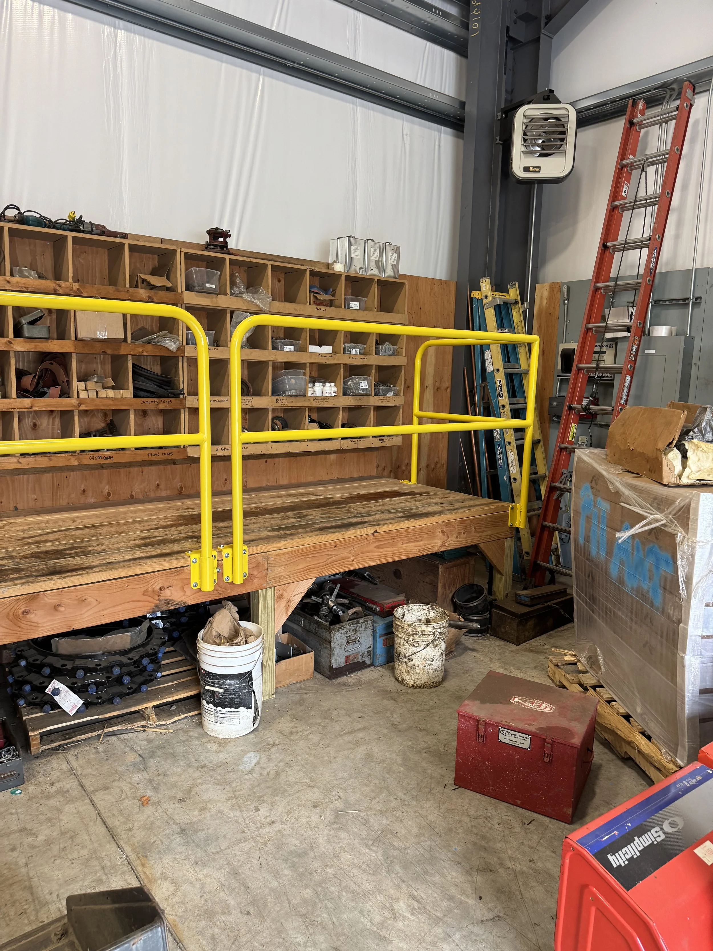 A workshop with a wooden workbench, yellow safety rails, shelving with small parts, and ladders leaning against the wall.