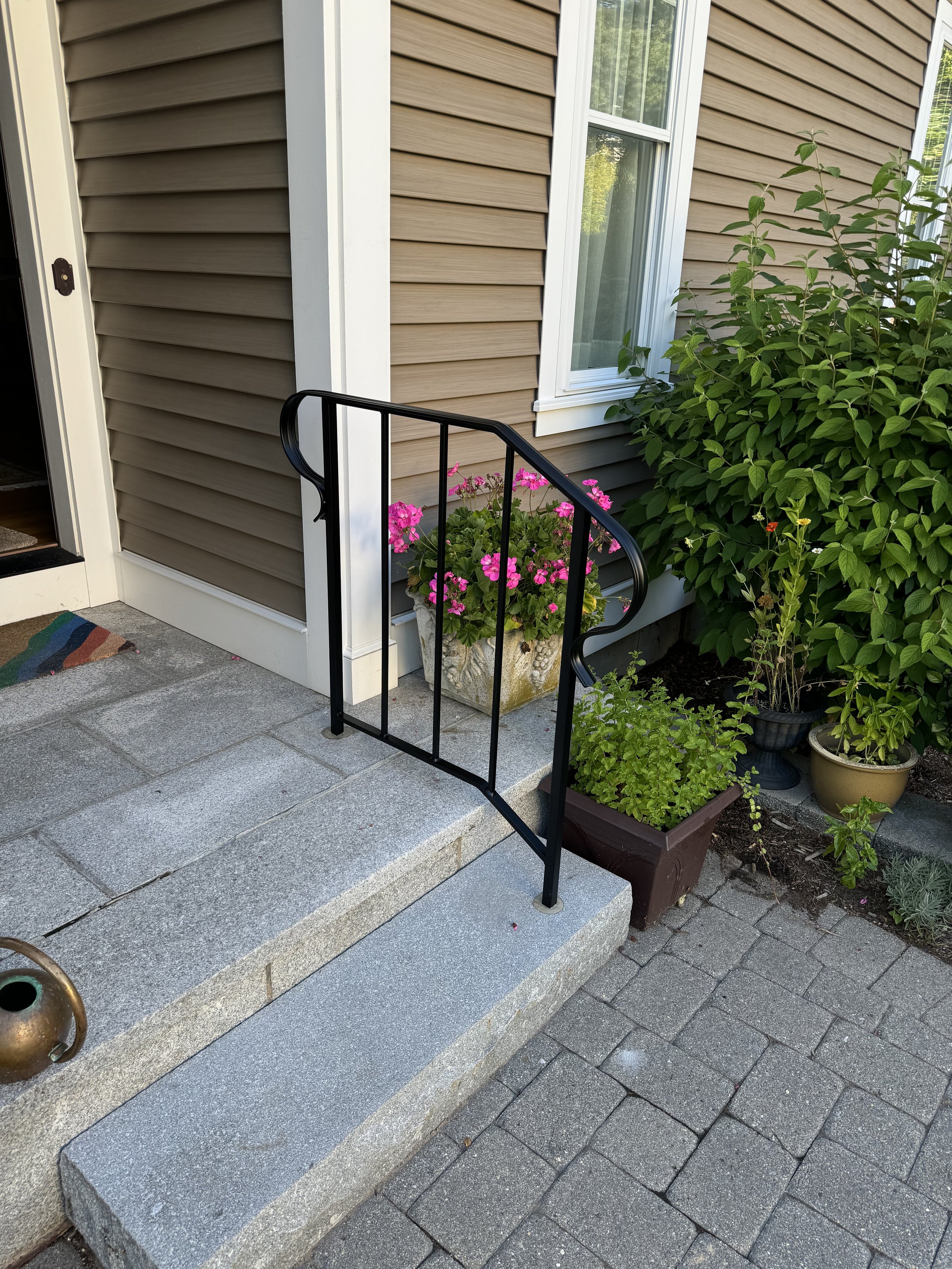 Front porch with stone steps, black metal railing, potted pink flowers, and green plants next to a house with beige siding and a white-framed window.