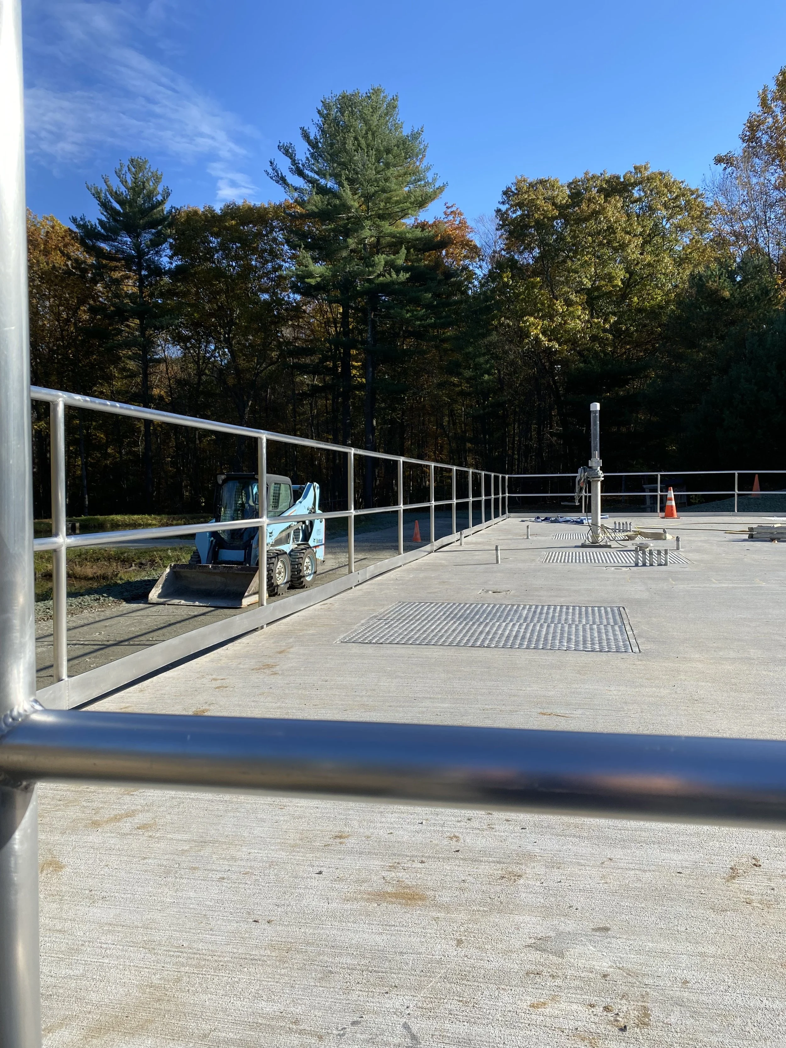 An empty concrete rooftop with a small blue construction vehicle, metal railings, and trees in the background under a partly cloudy blue sky.