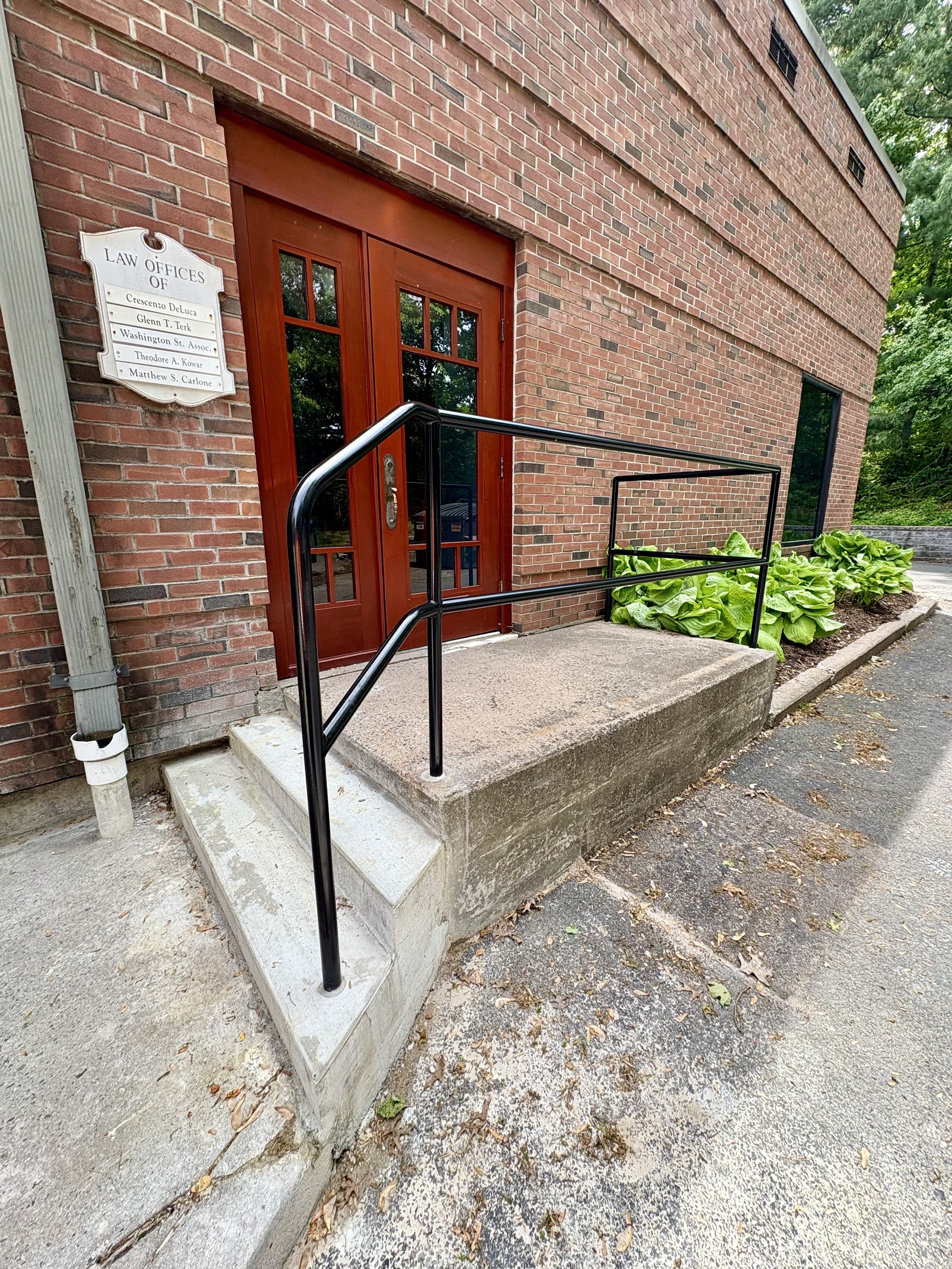 Red brick building with a wooden door, a small concrete ramp with black metal railing, and a sign indicating law offices. There are leafy green plants beside the ramp.
