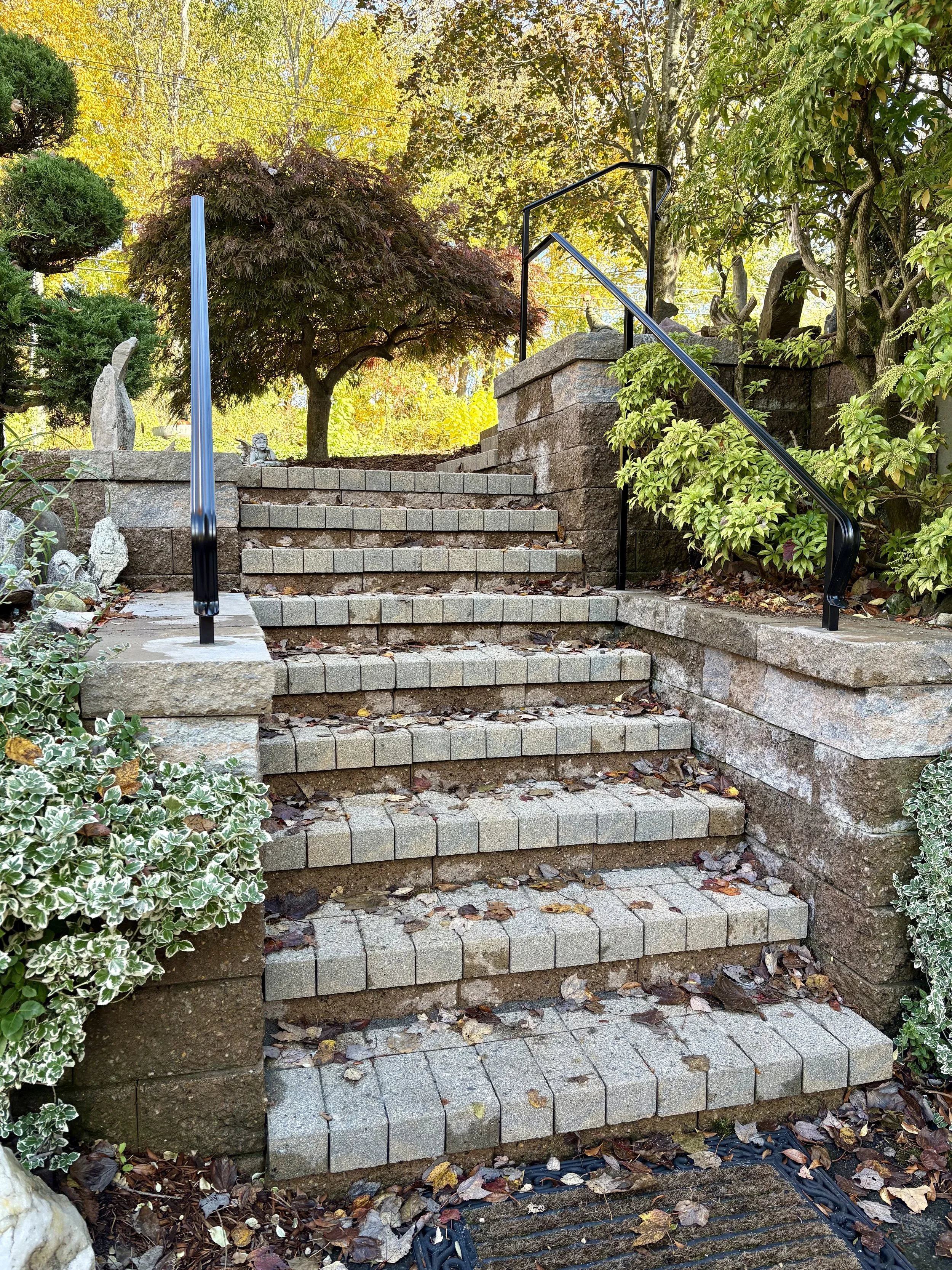Stone outdoor staircase with leaves scattered on steps, surrounded by greenery and trees, with a black handrail on the right side.