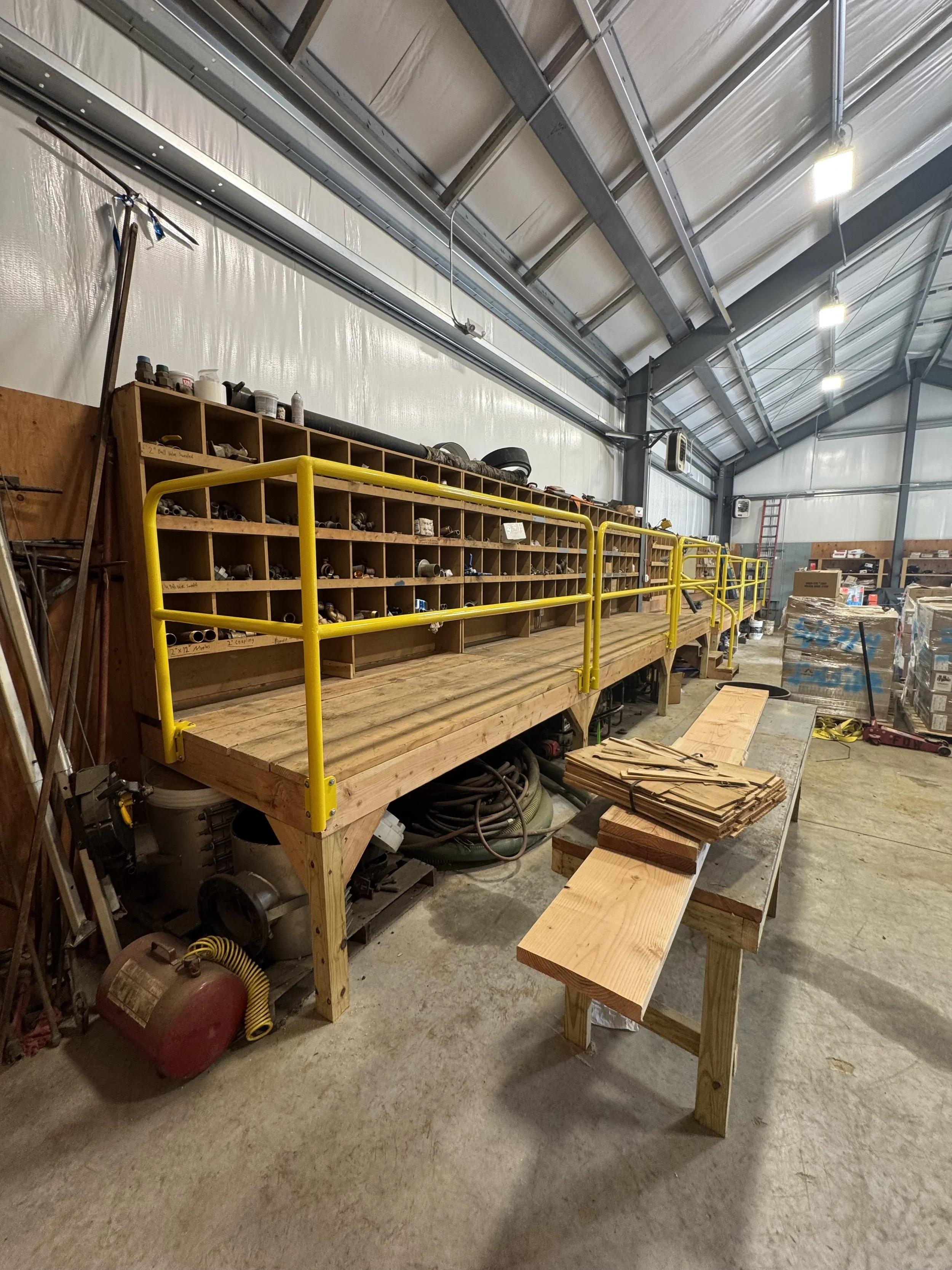View of an organized woodworking workshop with a wooden workbench, yellow safety rails, and shelves filled with small parts and tools, in a large industrial space.