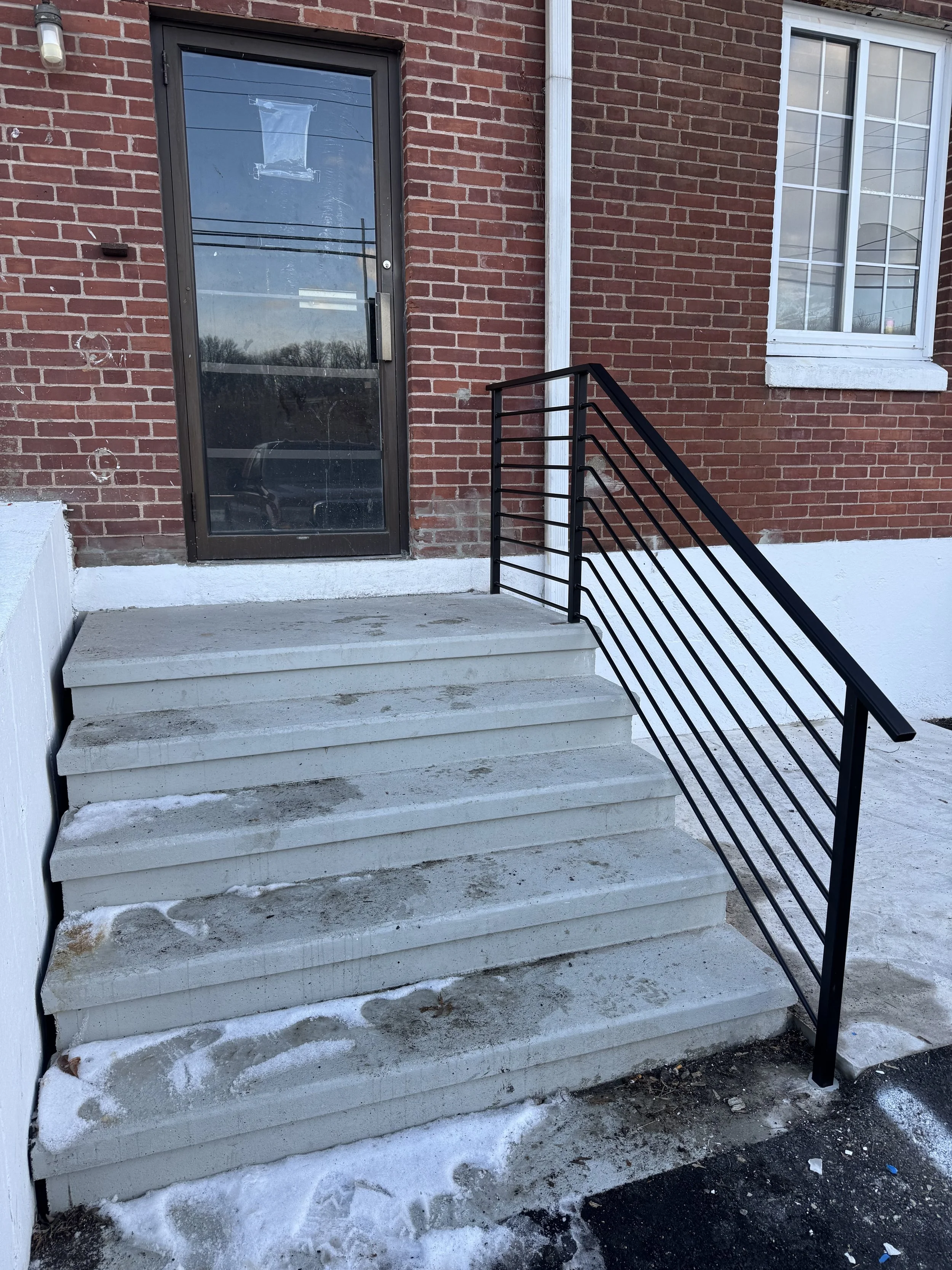 Concrete stairs with a black metal handrail leading to a glass door with a brick exterior wall and a large window nearby.