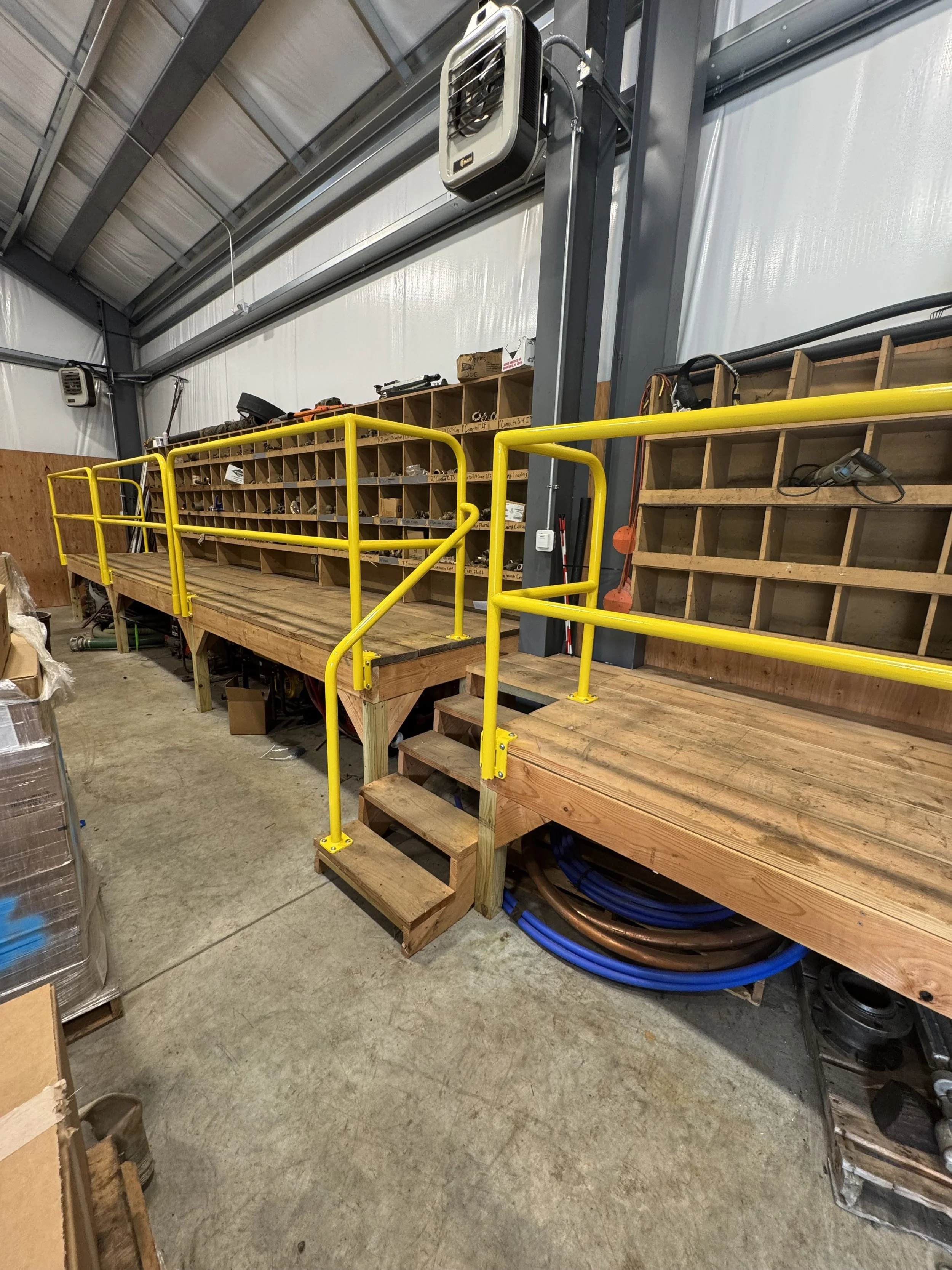 A wooden industrial workspace platform with yellow safety railings and stairs inside a warehouse, with storage cubbies and tools.