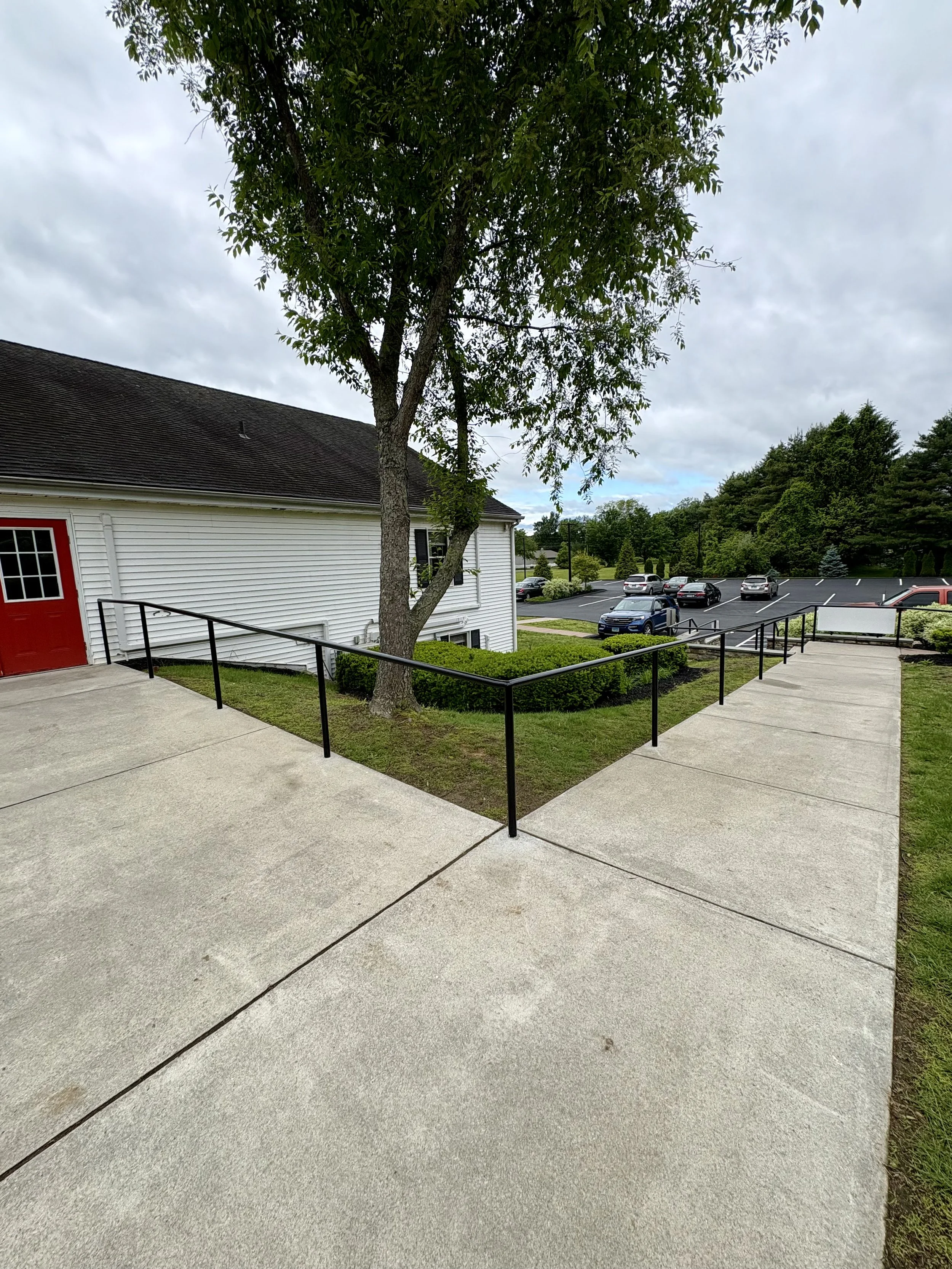 Concrete sidewalk with black metal handrails leading around a large tree near a white building with a red door, parking lot with parked cars, grassy areas, and trees in the background under a cloudy sky.