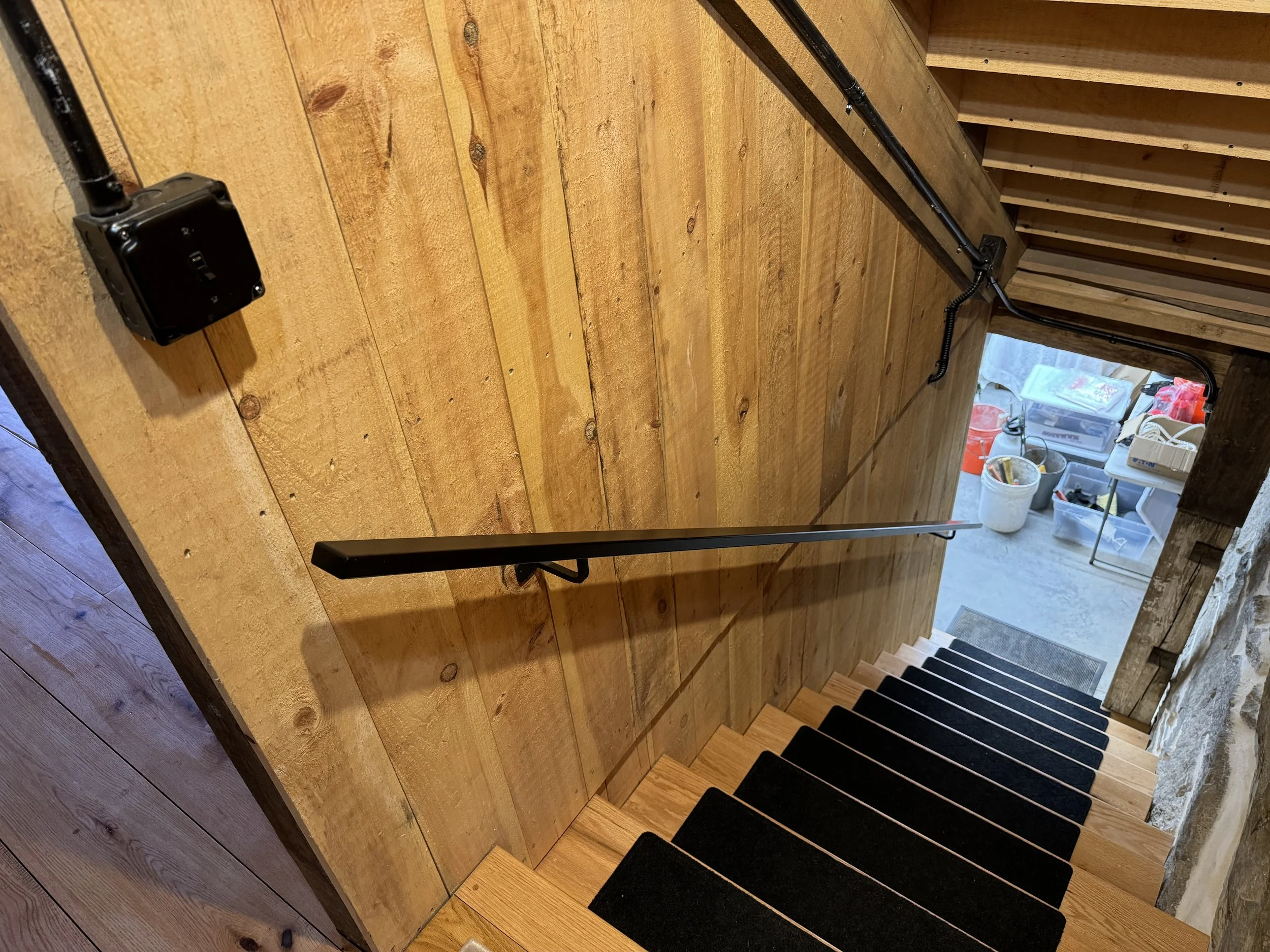 View looking down a wooden staircase with a black handrail. The stairs have black mats on each step. At the bottom, there are storage containers, buckets, and various tools and items in a garage or workshop area.