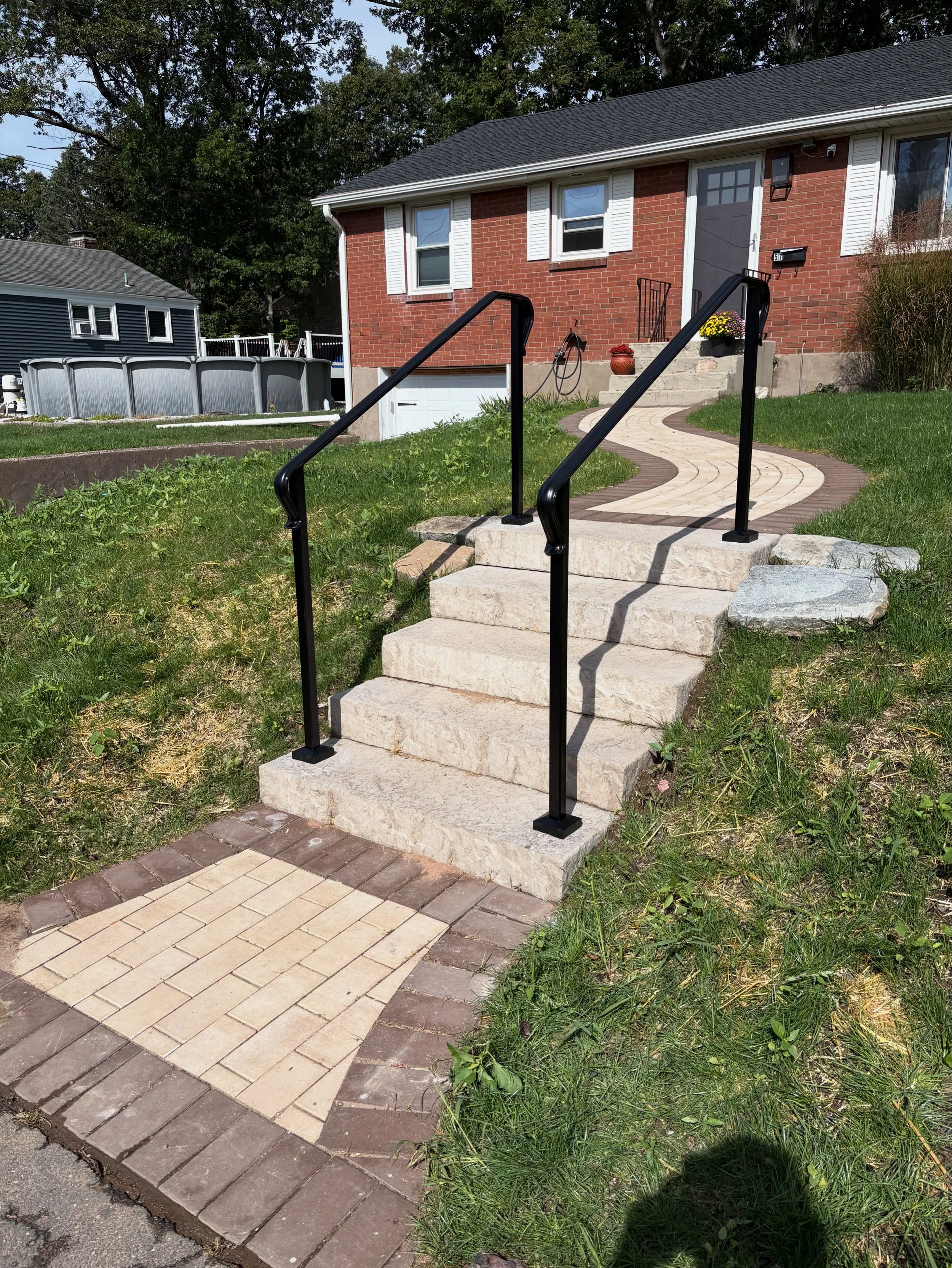 Stone stairs with black metal handrails leading up to a front door of a brick house with white trim and a small porch. There is a paved walkway, grass, and a large rock to the side.