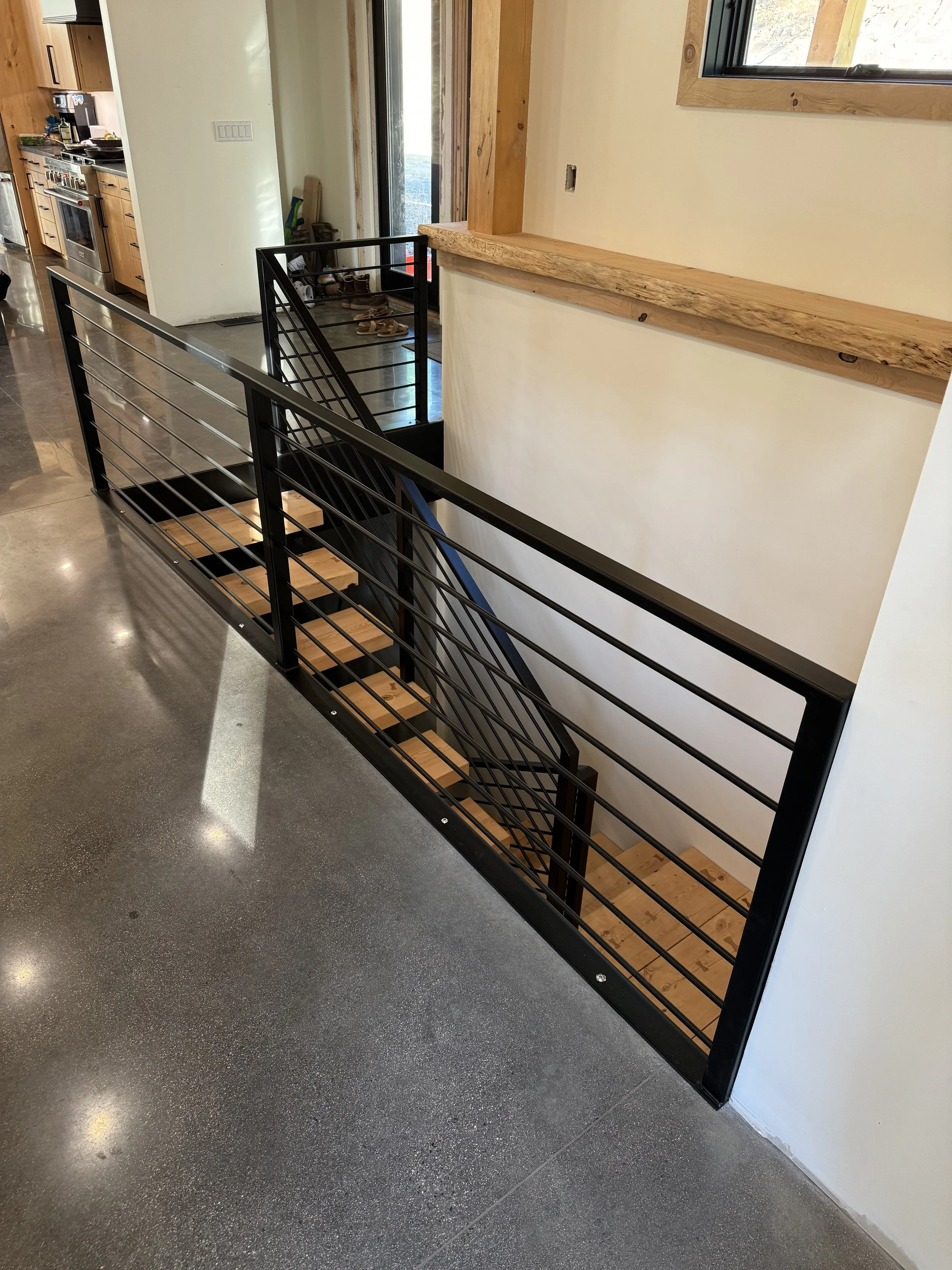 Interior view of a modern home with a staircase featuring a black metal railing and wooden steps, next to a wall with a wooden ledge, and a kitchen area in the background with appliances and cabinets.