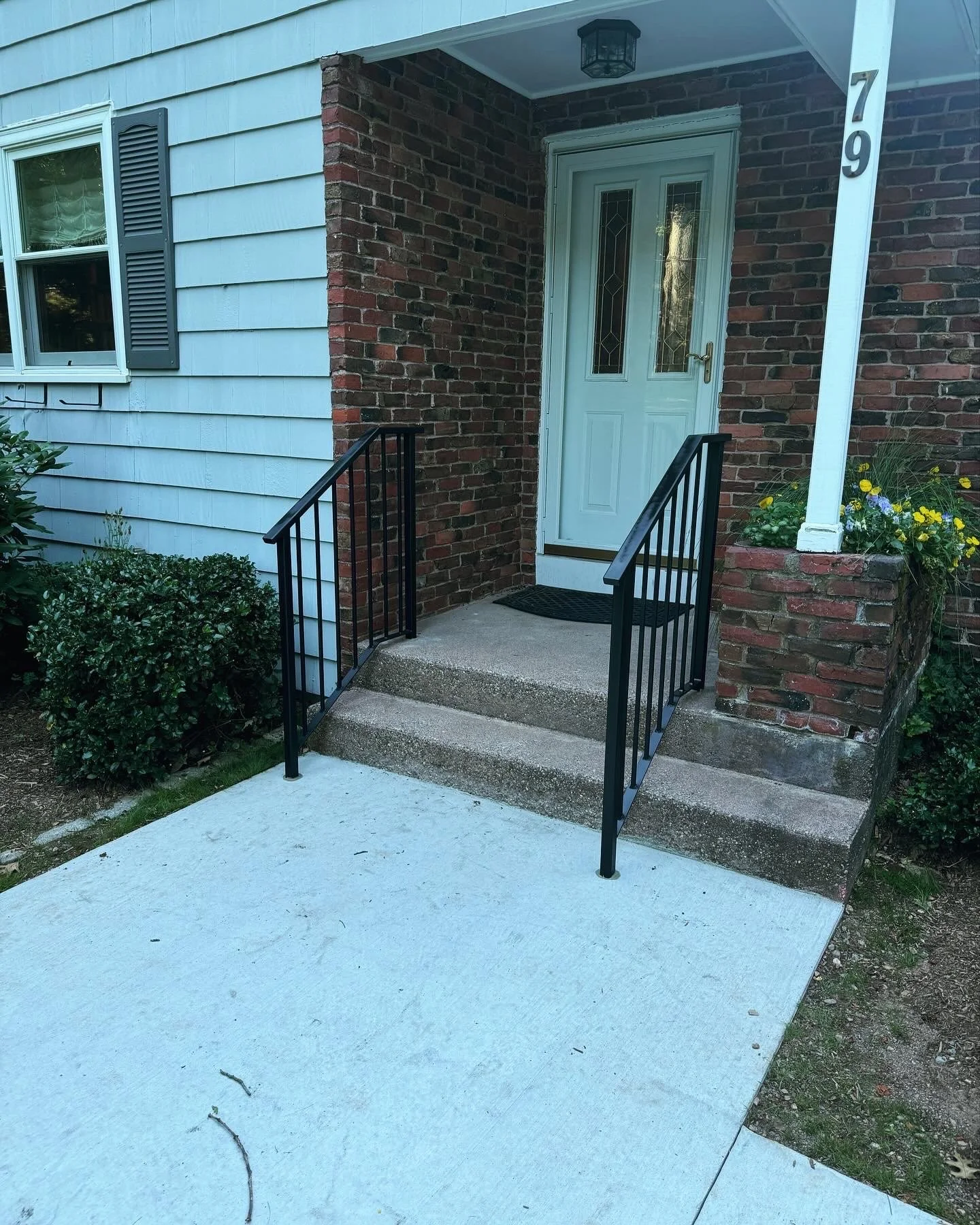 Front porch with stairs, black metal handrails, white door with window panels, brick and board exterior wall, flower bed with yellow and purple flowers, concrete sidewalk in front of the house.