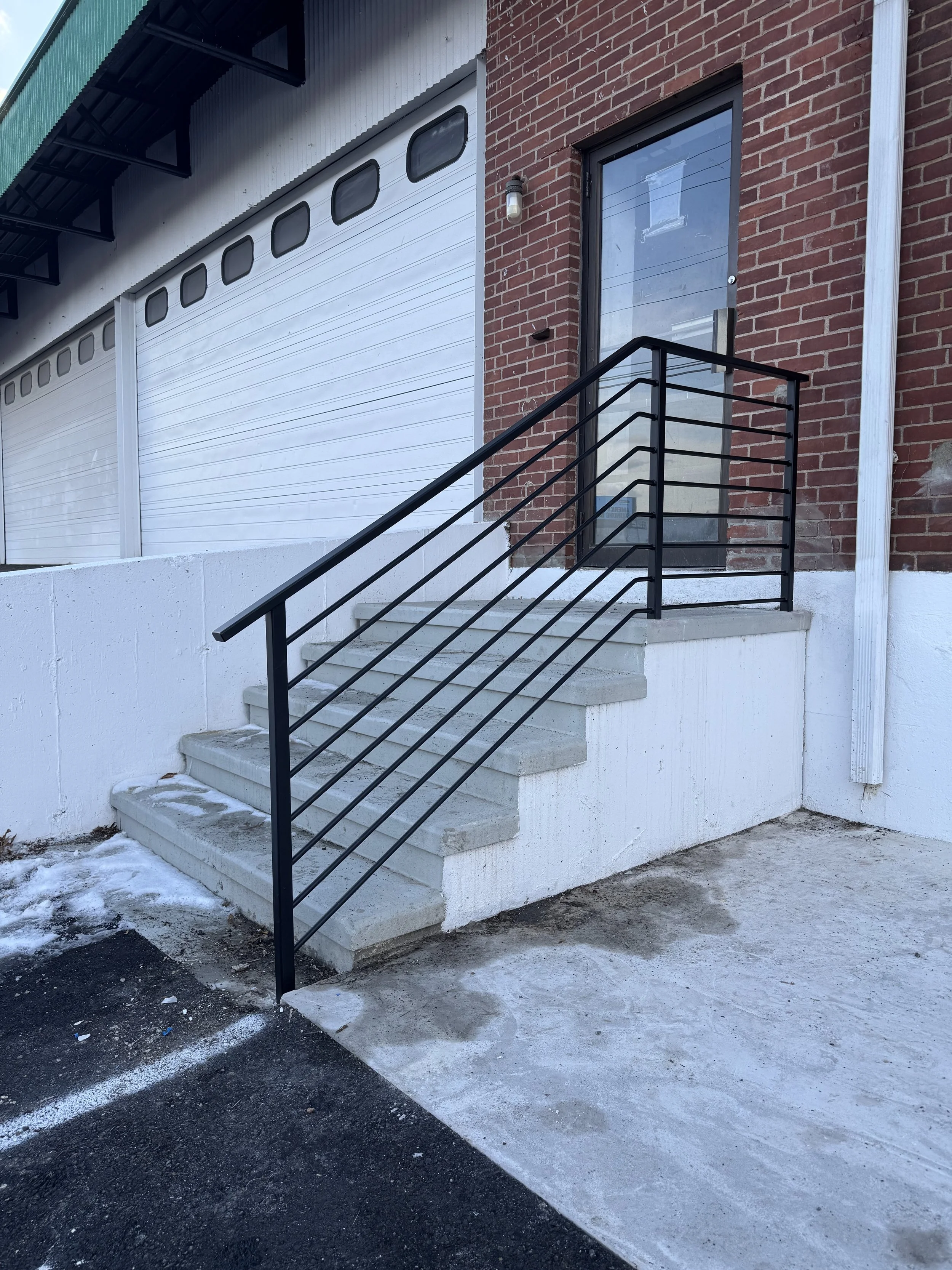 Concrete stairs with black metal railing leading to a glass door on a brick building, snow on the ground.