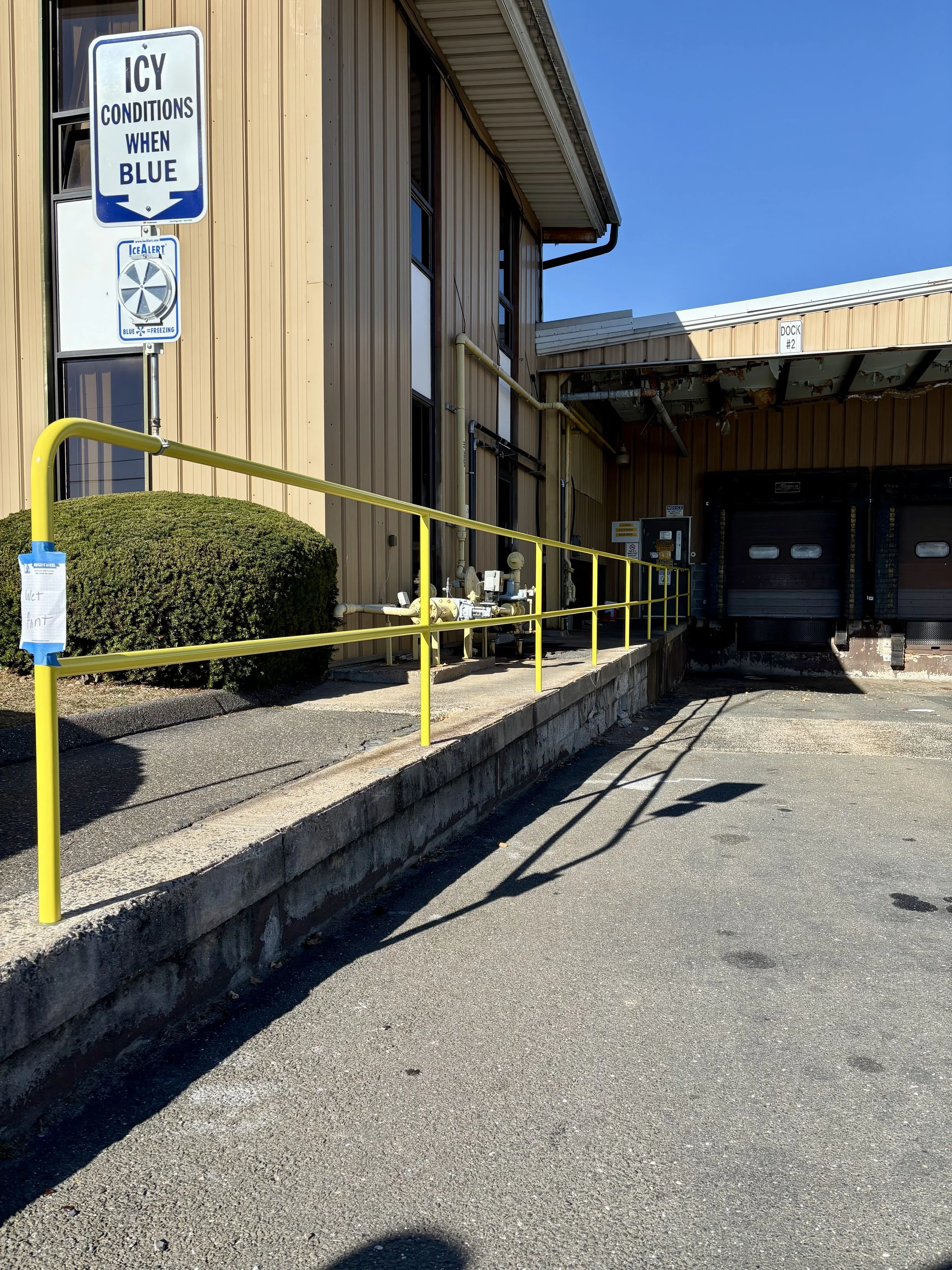 Exterior of a building with a loading dock, yellow railing, sign indicating icy conditions when blue, and a bush next to the wall.