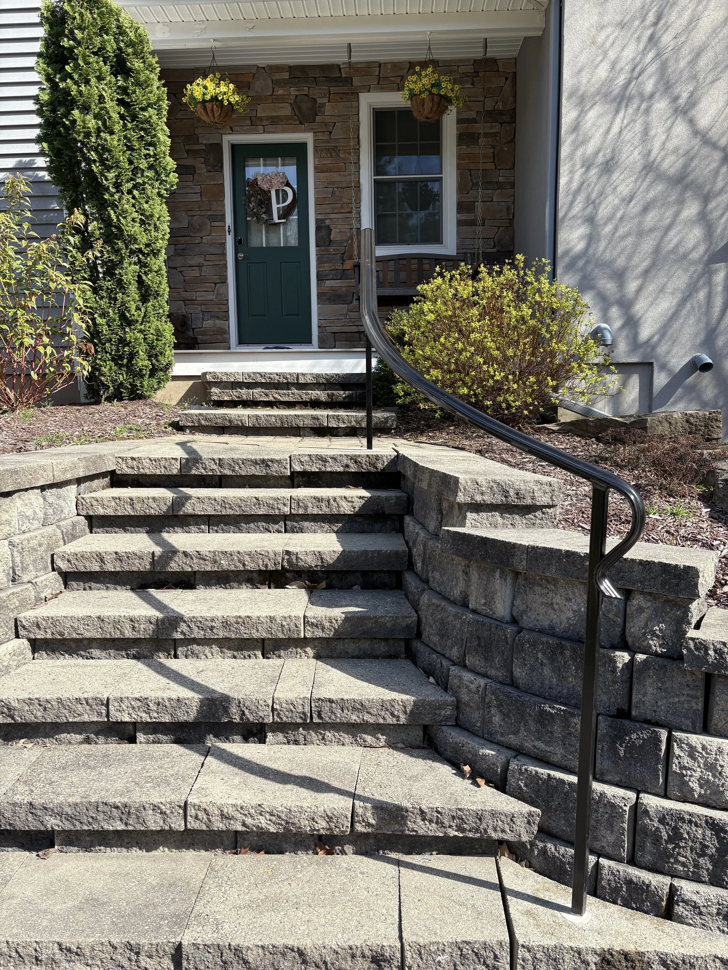 Stone steps leading to a porch with a green front door, surrounded by shrubs and plants, with hanging flower baskets above.