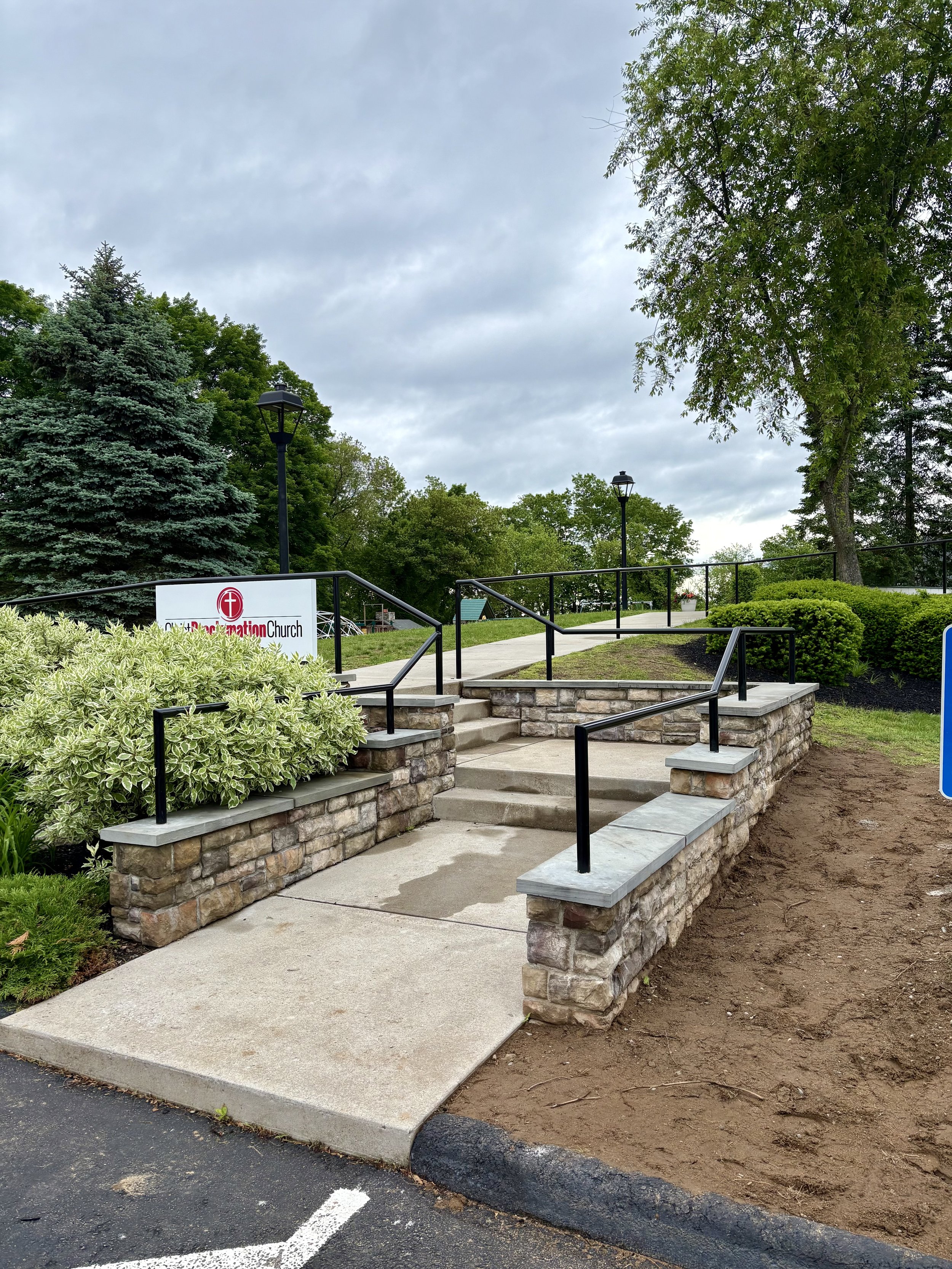 A wheelchair accessible ramp with metal handrails in front of a church, with trees and cloudy sky in the background.