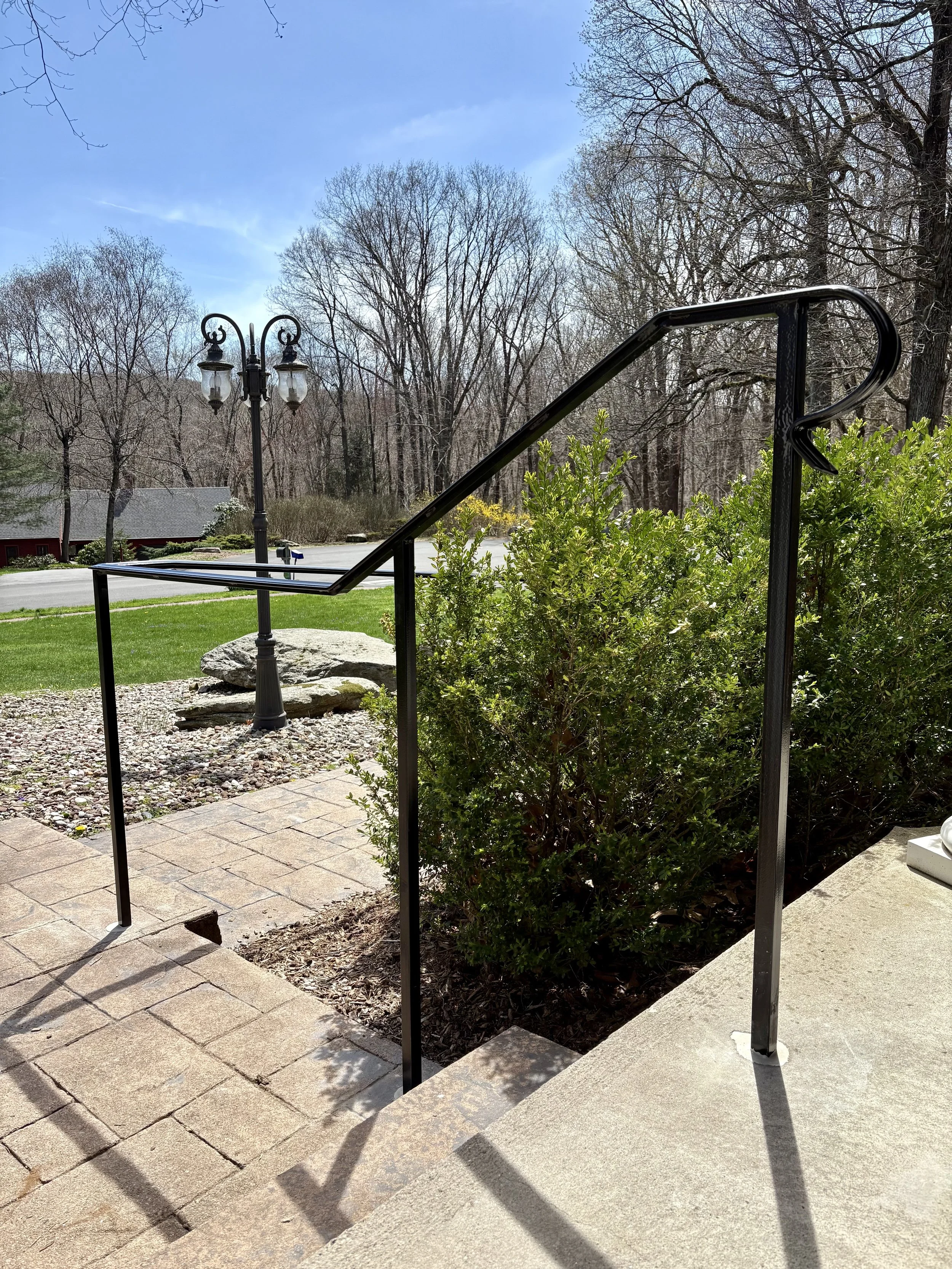 Black metal railing on concrete steps leading down to a garden with a bush. In the background, there is a lamppost, a grassy yard, trees, a house, and a street with a mailbox, under a partly cloudy blue sky.