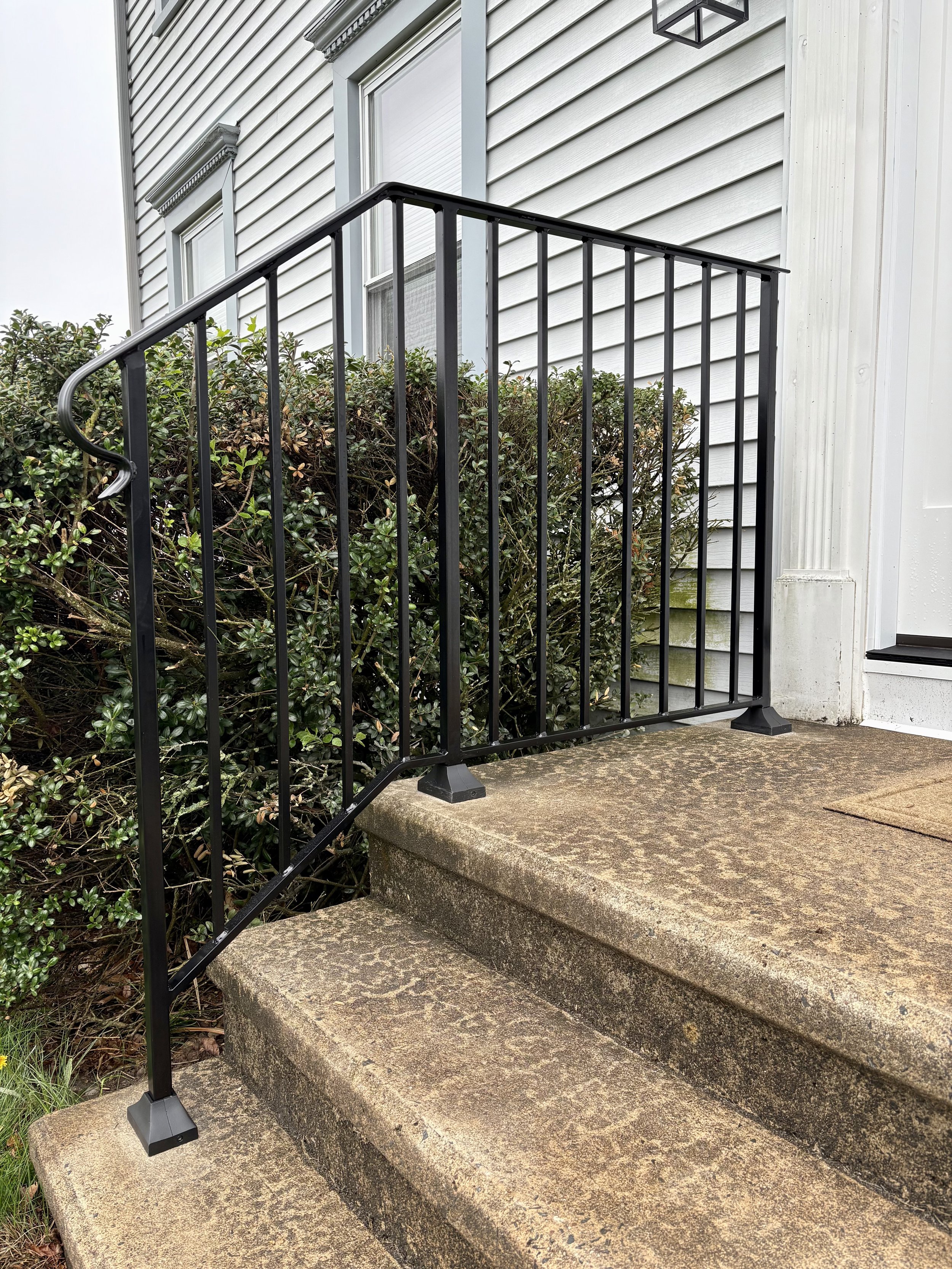 Black metal railing on stone steps leading to a white house with beige siding and windows, a bush on the side, and a black outdoor light fixture.