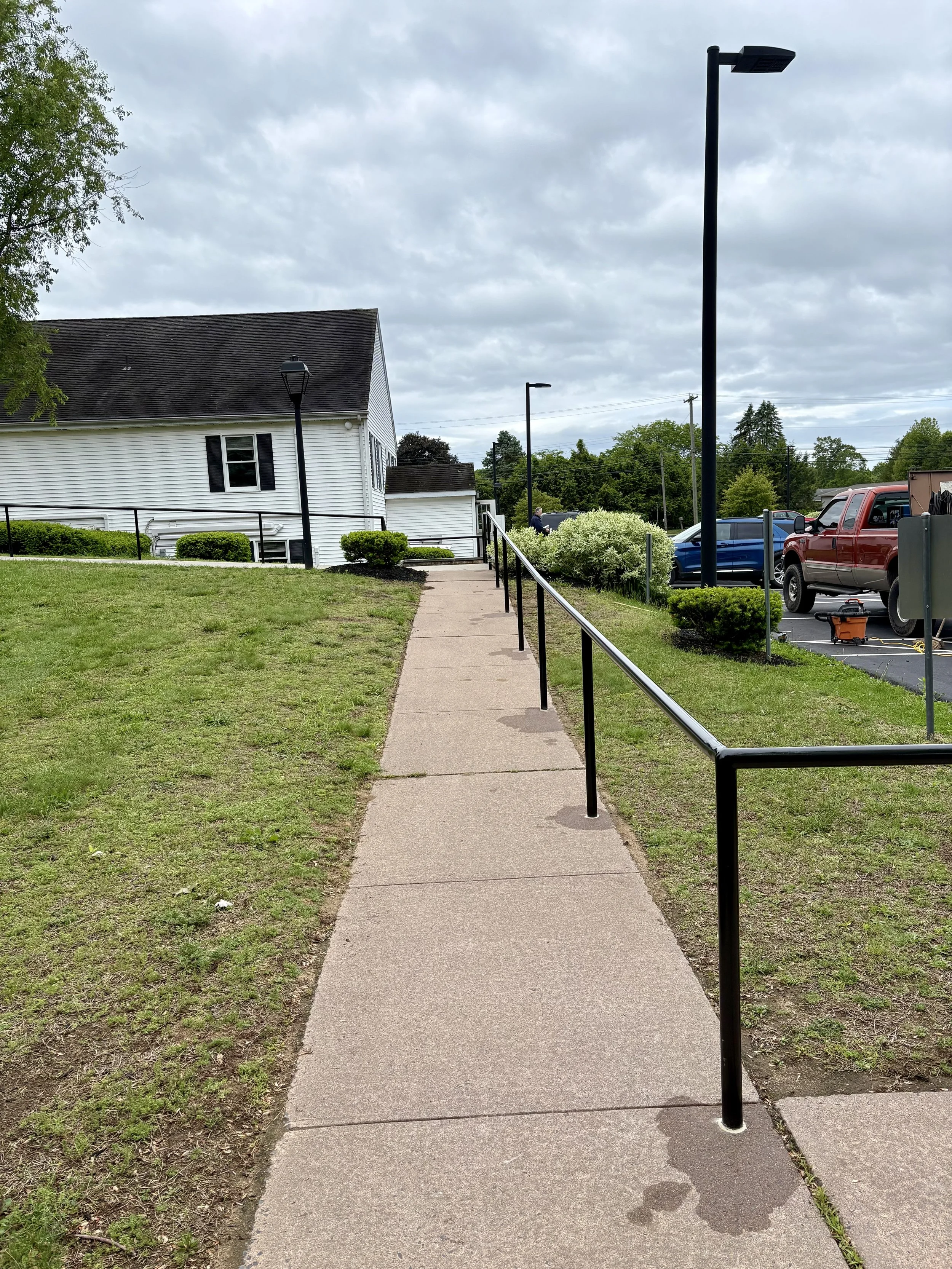 A sidewalk with a black railing on the right side, leading uphill to a white house with black shutters. There is a grassy area on the left and a parking lot with cars on the right. Overcast sky with gray clouds.