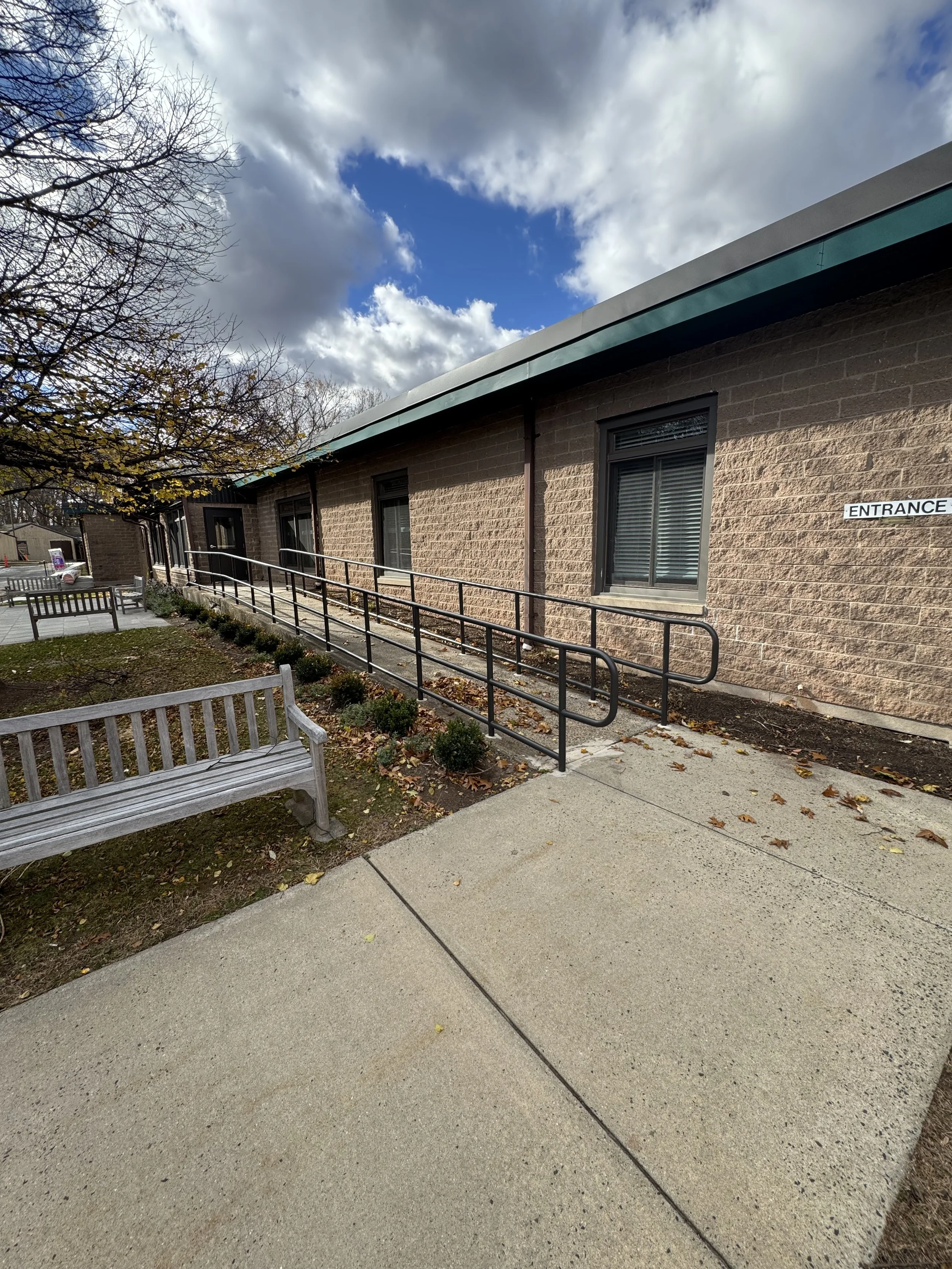 Exterior view of a brick building with parking lot and benches, featuring a wheelchair ramp with black railings and a sign that says 'ENTRANCE'.