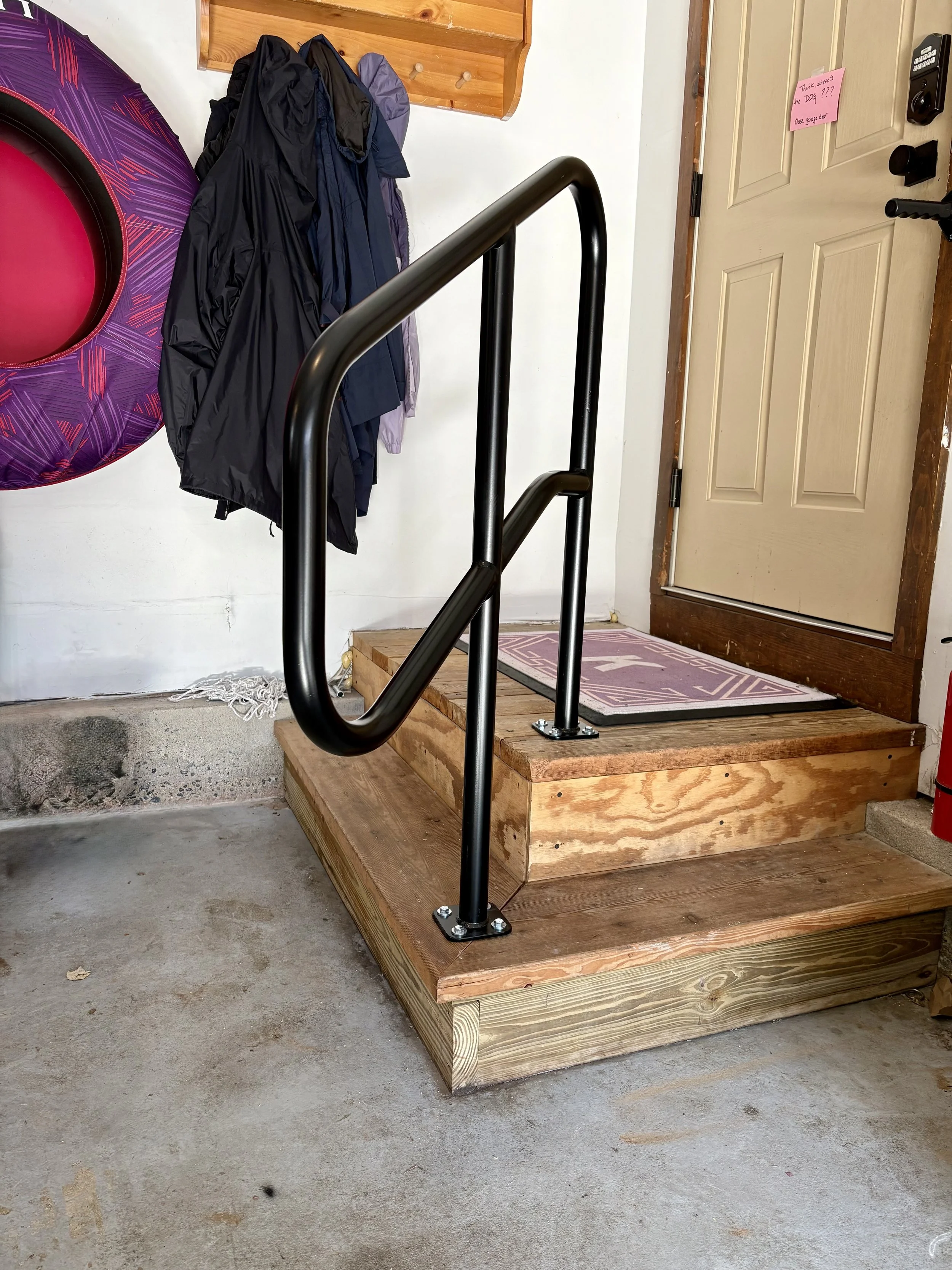 Wooden staircase leading to a front door with a pink welcome mat and a black handrail, in a garage with jackets hanging on the wall and a purple inflatable ring.