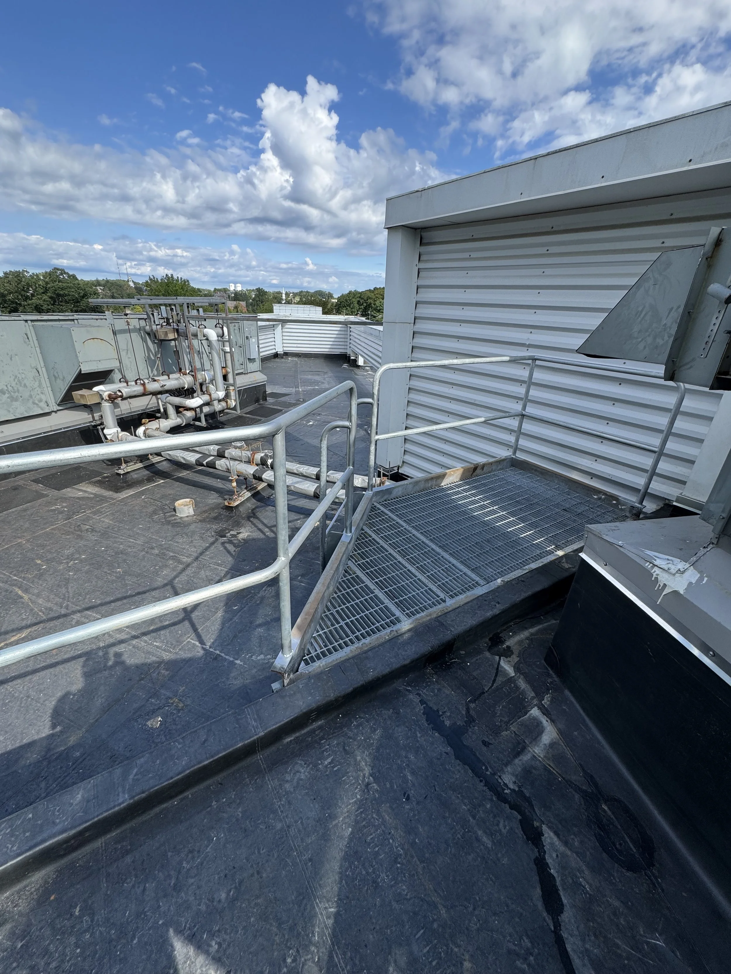 Rooftop with metal railing and grated drainage cover, HVAC equipment, and white vinyl siding, under a partly cloudy blue sky.