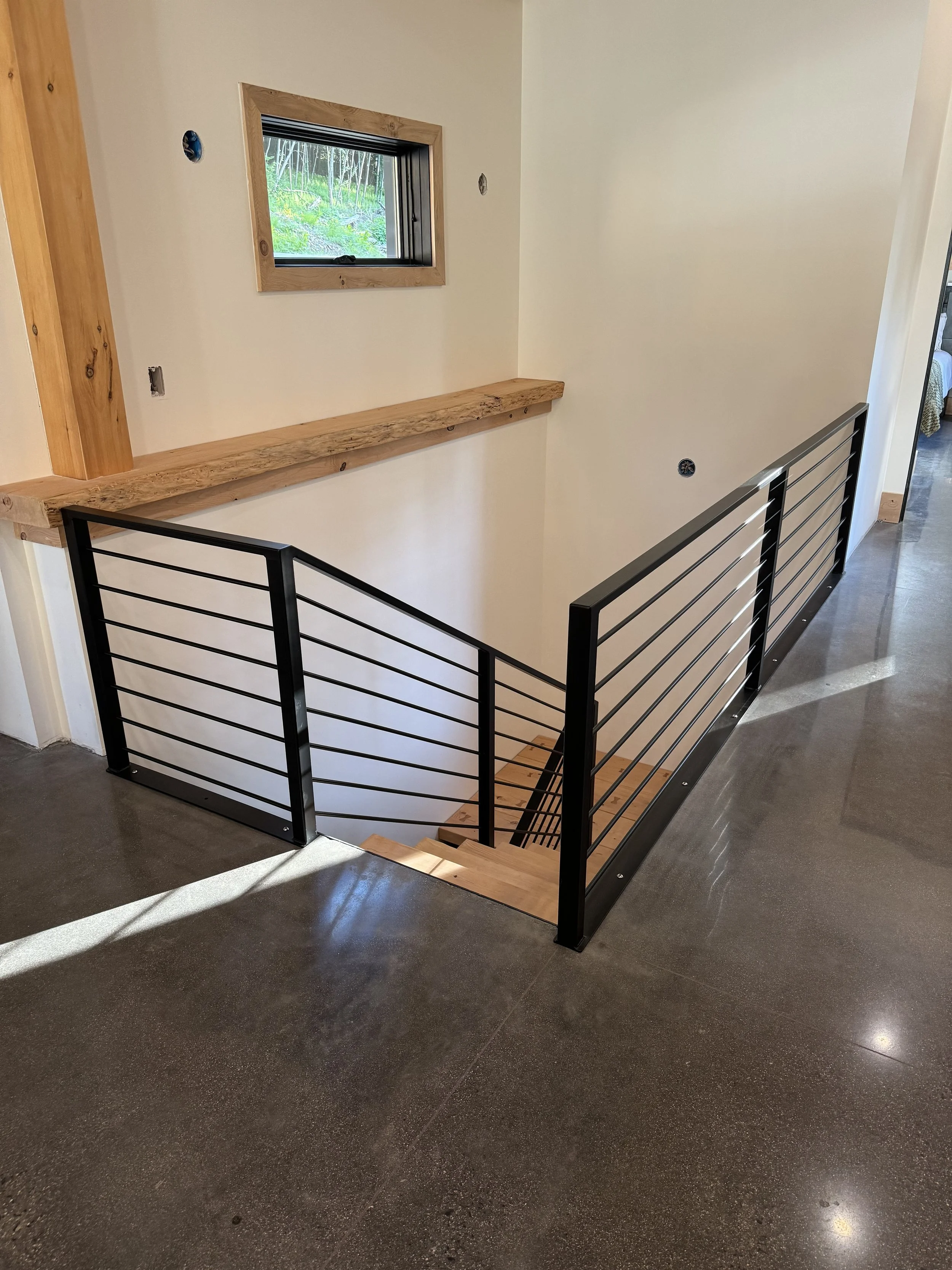 Interior view of a staircase with black metal railing, wooden steps, a wooden handrail, and a small window showing greenery outside.
