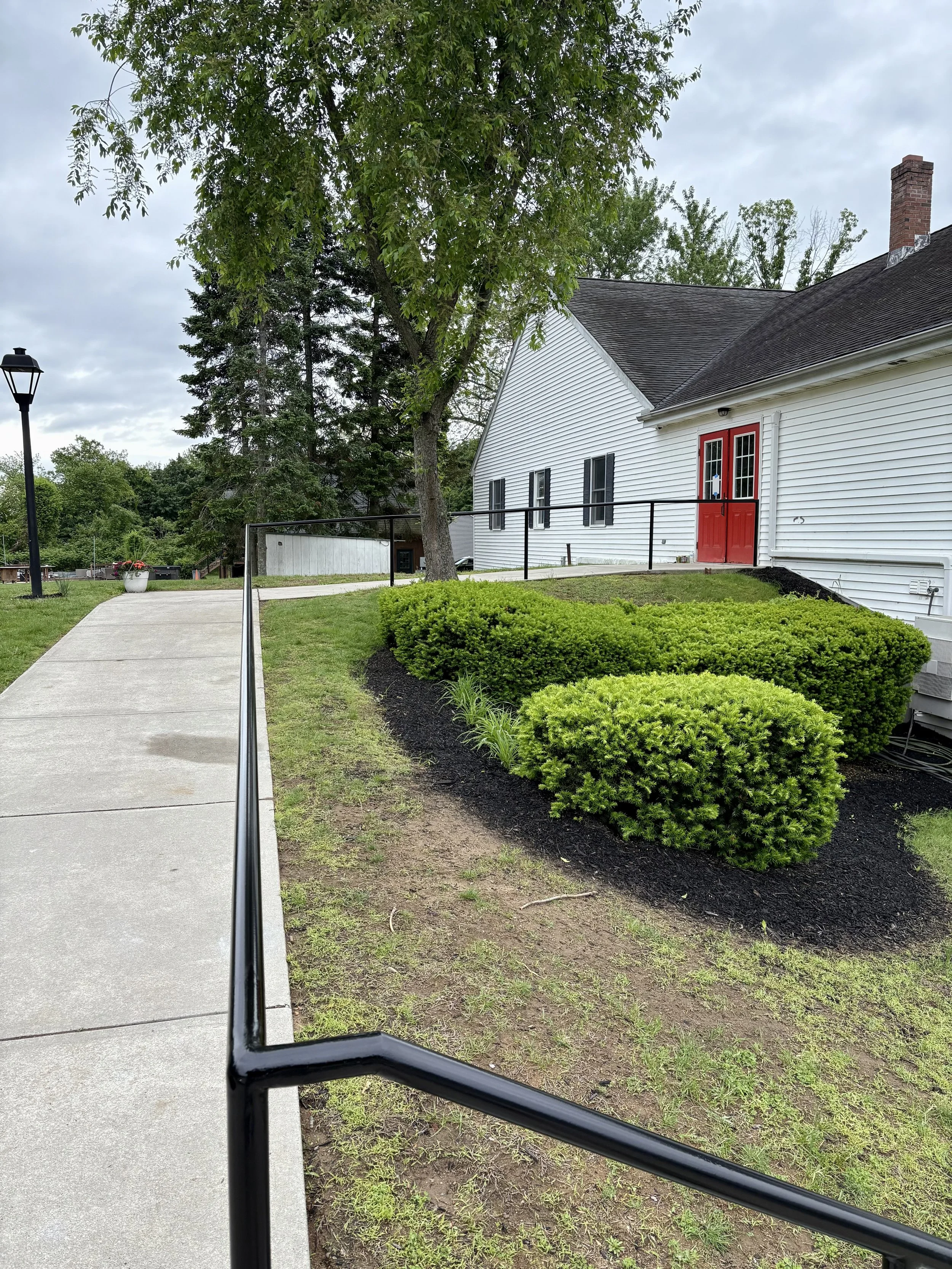 White building with a red door, surrounded by neatly trimmed bushes and a concrete walkway with a black metal handrail, in a green outdoor area with trees and a cloudy sky.