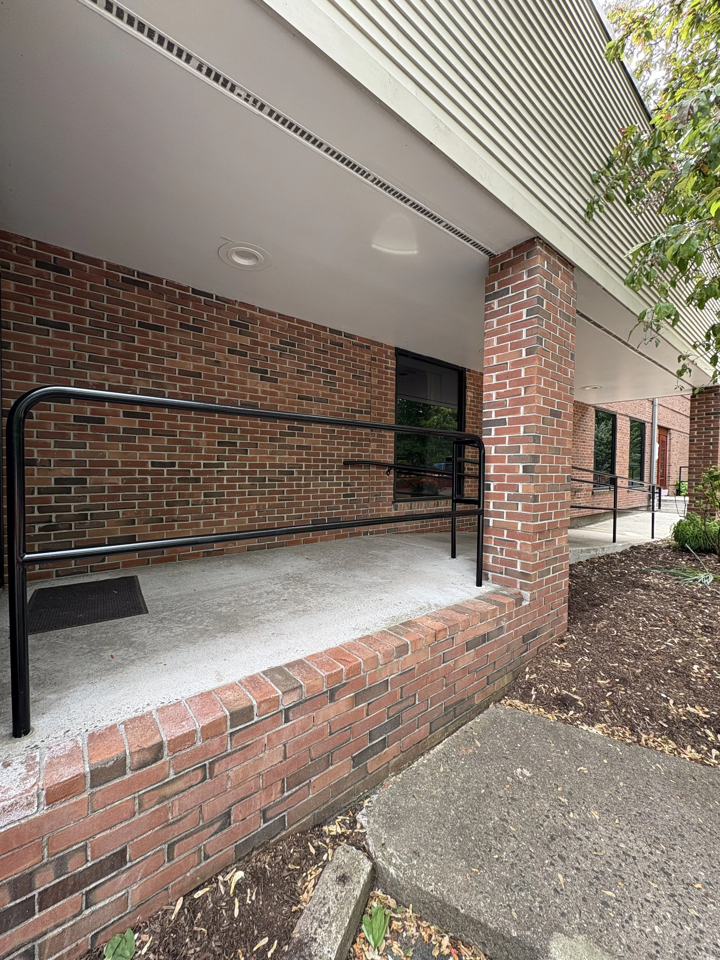 A brick building entrance with a concrete ramp and black metal handrails, surrounded by soil and some greenery.