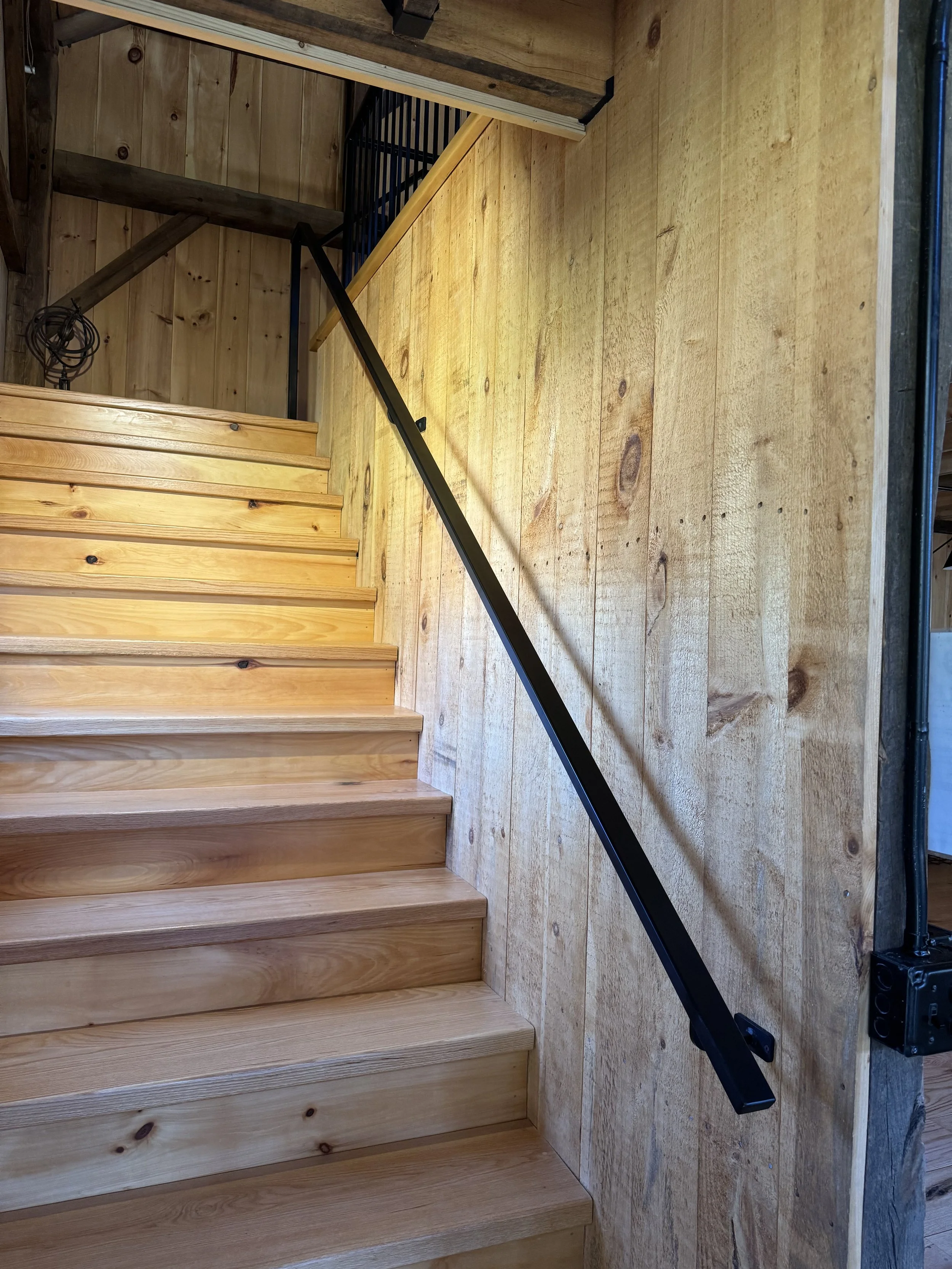 Wooden staircase with a black metal handrail on a wood-paneled wall in a rustic interior.