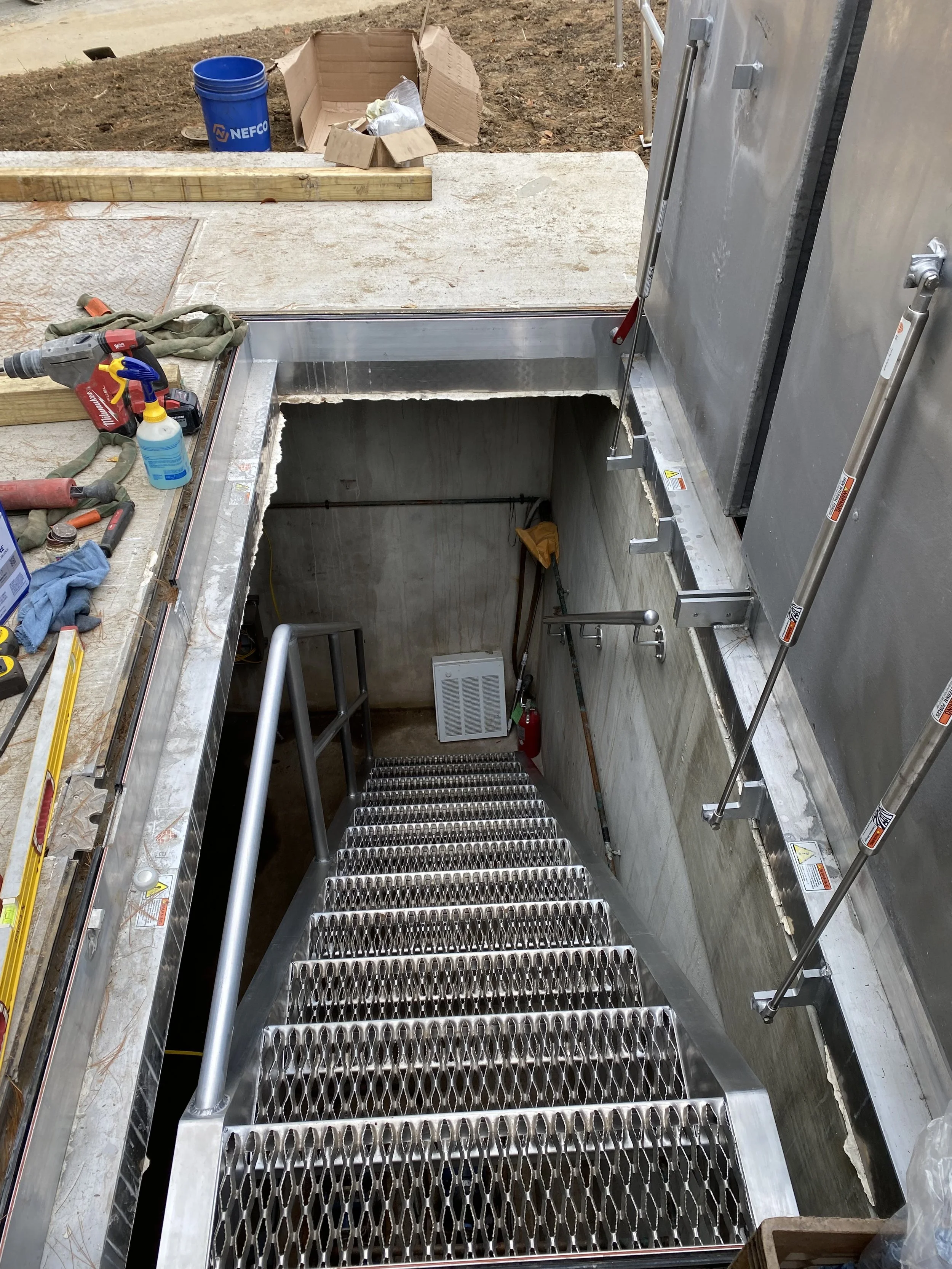 Construction site showing a metal staircase leading down to an underground space, with construction tools and materials on the surrounding surface.