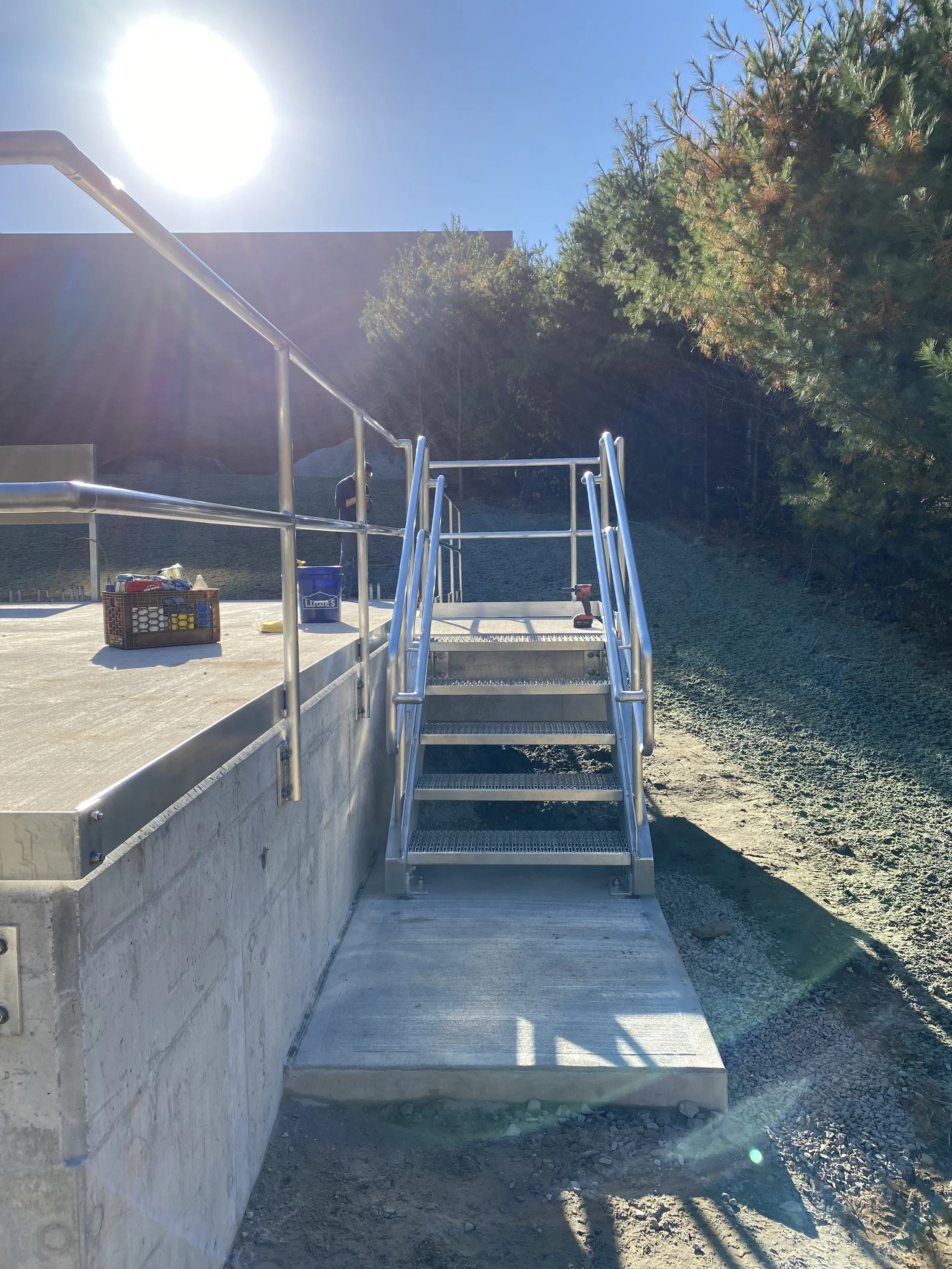 Metal platform with stairs and handrails, outdoors with trees and a building in the background, sunlight bright in the sky.