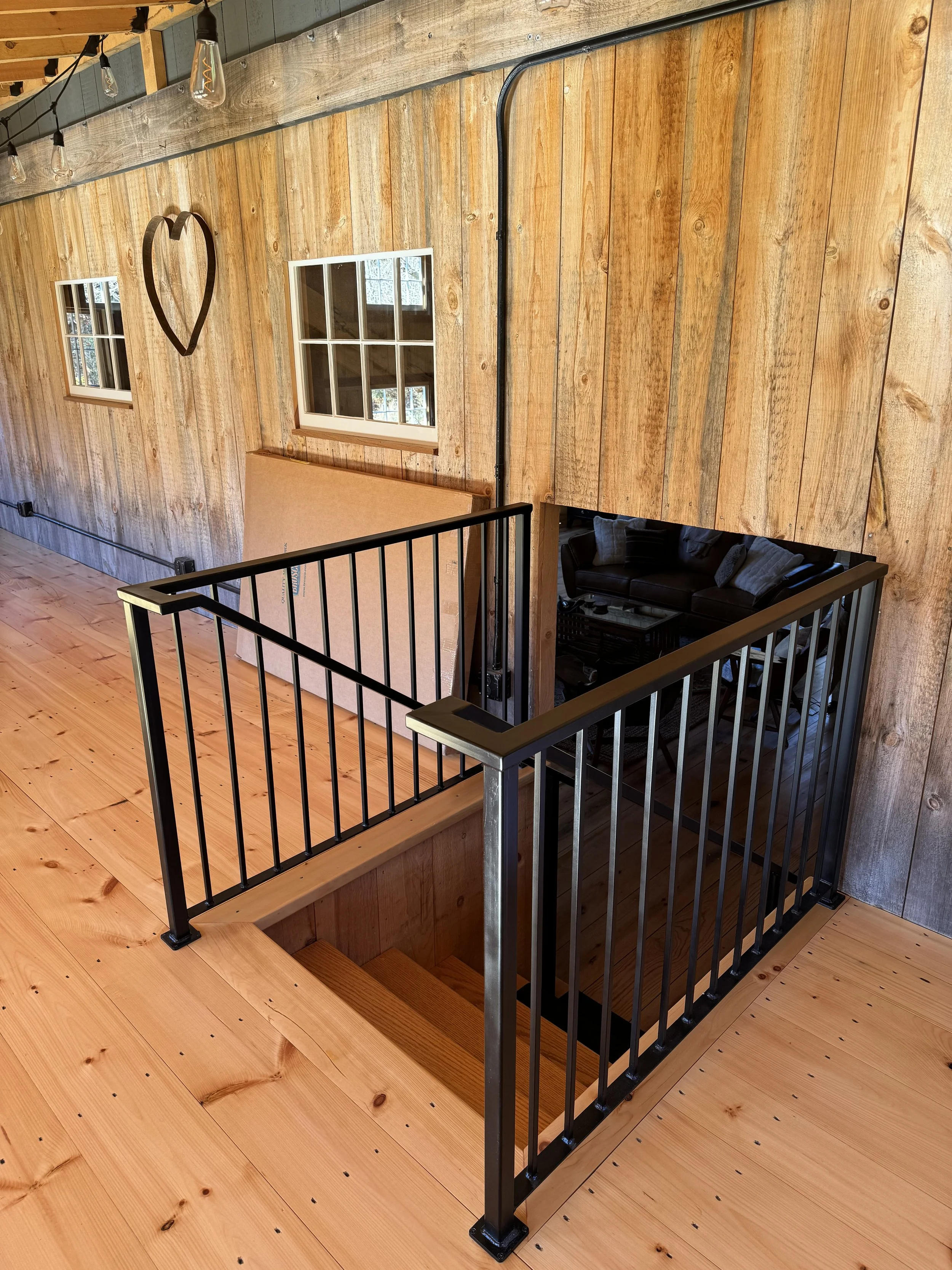 Interior view of a wooden house featuring a staircase with a black metal railing, wooden walls and flooring, two small square windows, a heart-shaped wall decoration, and framed artwork visible in a cozy living space below.