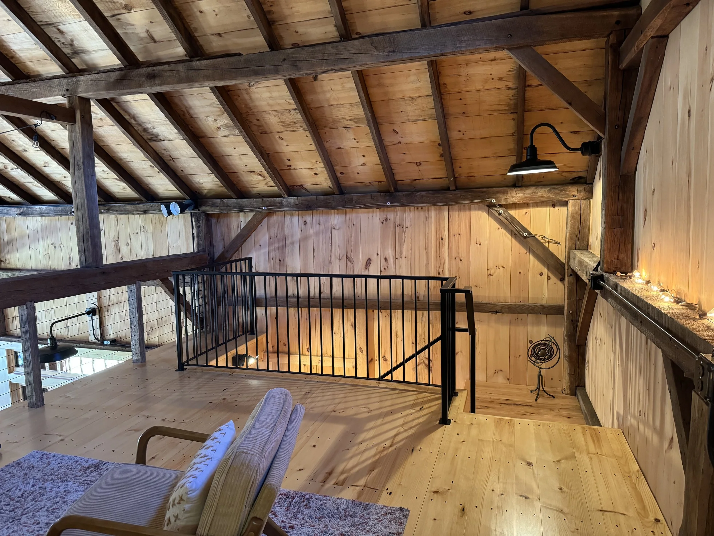 View of a cozy attic space with wooden walls, ceiling, and floor, illuminated by wall-mounted lights and string lights, with a chair and a throw pillow in the foreground, a black metal railing overlooking the staircase below.
