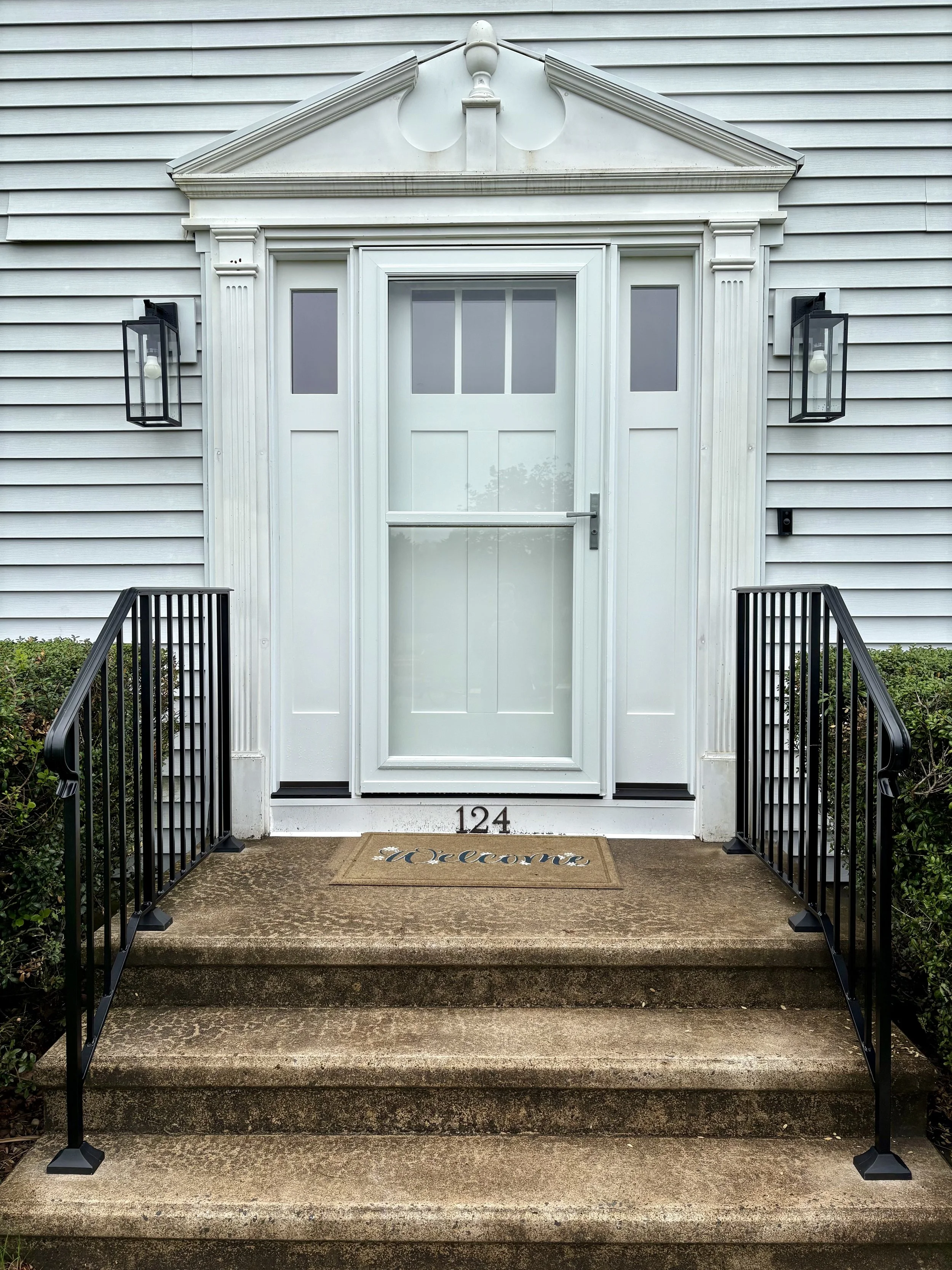 Front porch of a house with a white door, black metal railings, and a welcome mat, with the house number 124 visible.